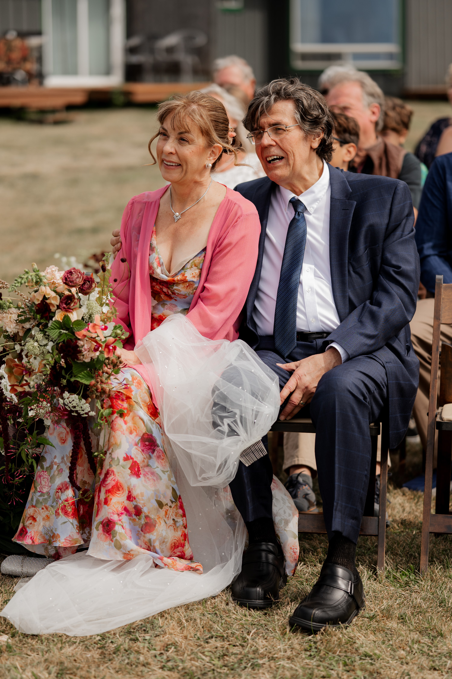 An elderly couple sits outdoors at a wedding, both wearing floral attire and holding a bouquet, smiling warmly in their wedding photos at the Yachats Inn.
