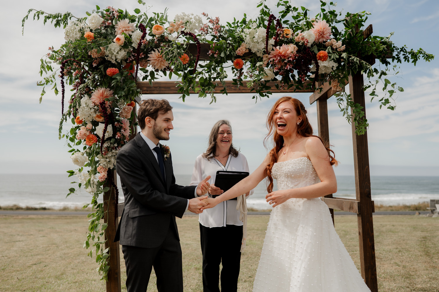 The bride and groom smile under a floral arch at an outdoor wedding ceremony by the sea, with the Yachats Inn as the venue.