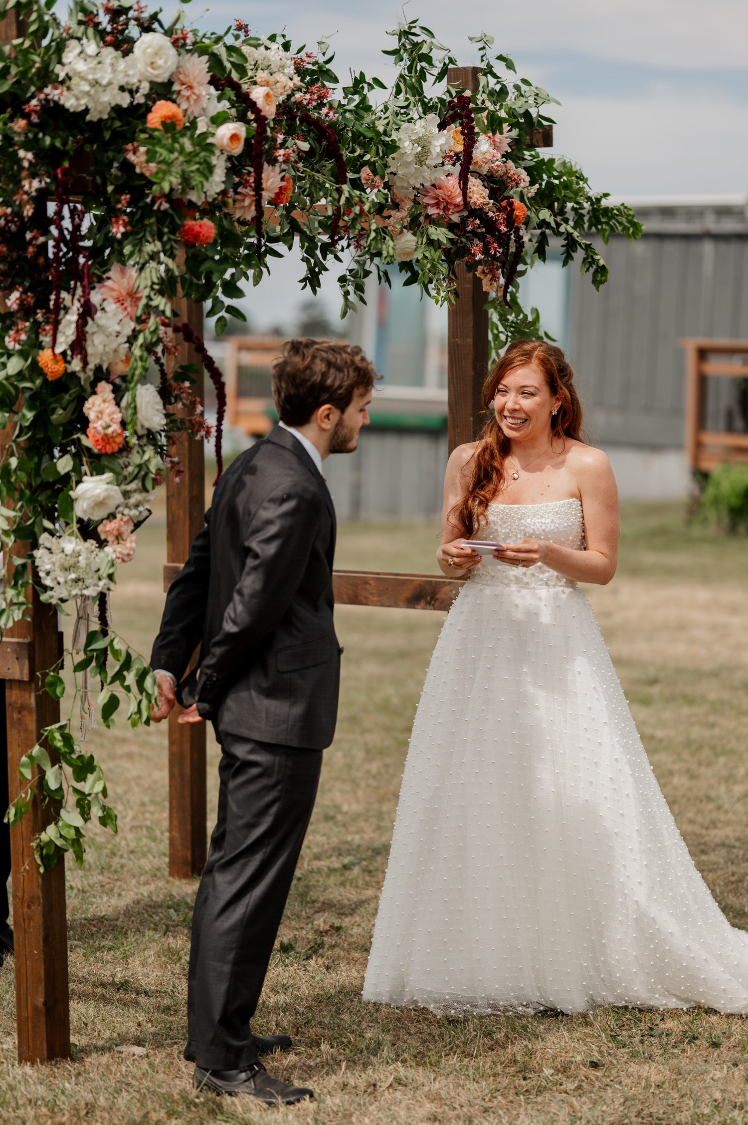 Bride and groom smiling under a floral arch in an outdoor setting at the Yachats Inn, with greenery and ocean visible in the background.