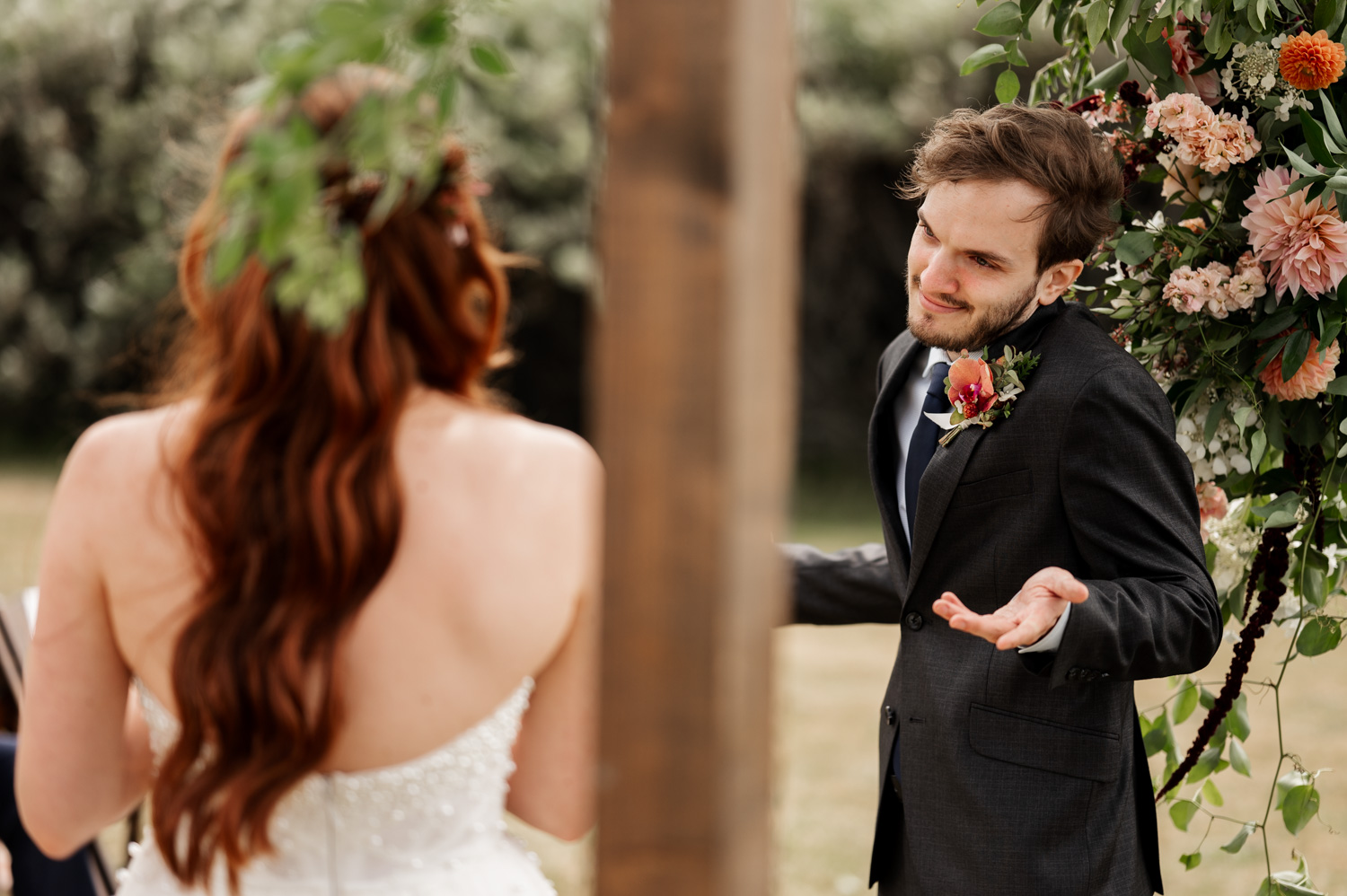 A groom playfully shrugs at his bride during an outdoor wedding ceremony at the Yachats Inn, with a backdrop of flowers adding to the whimsical moment.