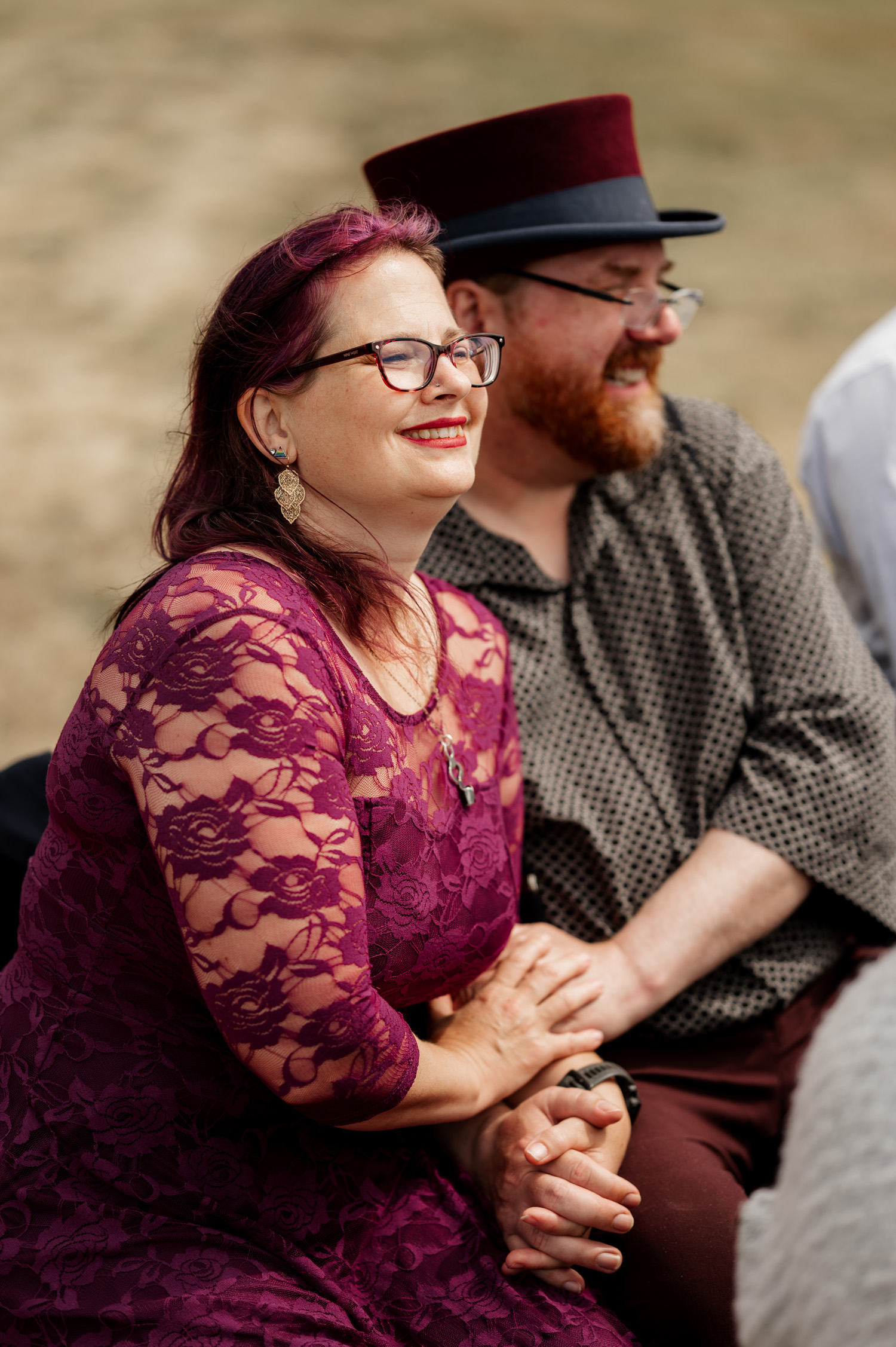 A woman in a purple lace dress smiles while holding hands with a man in a polka-dot shirt and top hat at the Yachats Inn. A scenic view of trees and gentle hills can be seen in the background, adding to the enchanting atmosphere of their wedding photos.