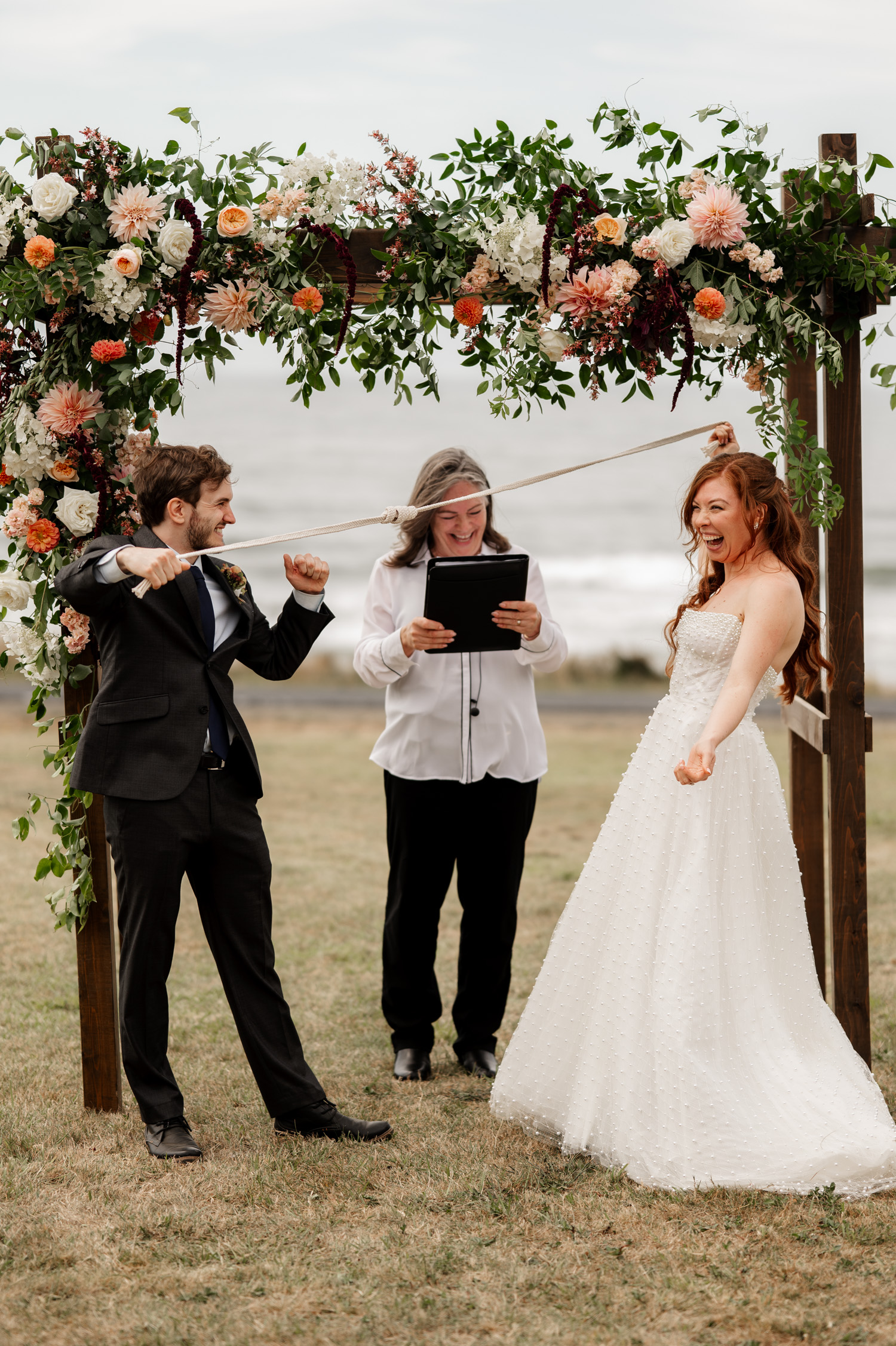 Bride and groom engage in a playful tug-of-war with a rope under a floral arch at the scenic Yachats Inn during their outdoor wedding ceremony.
