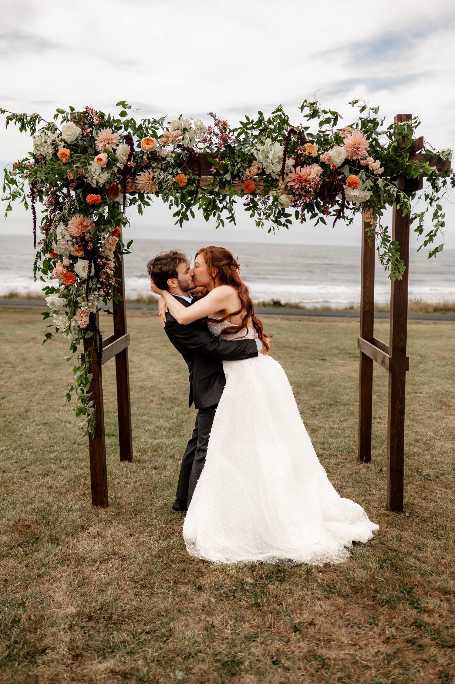 A couple shares a tender kiss under a floral arch by the ocean, with the bride in a white dress and the groom in a dark suit. The backdrop features waves gently crashing against the shore at Yachats Inn.