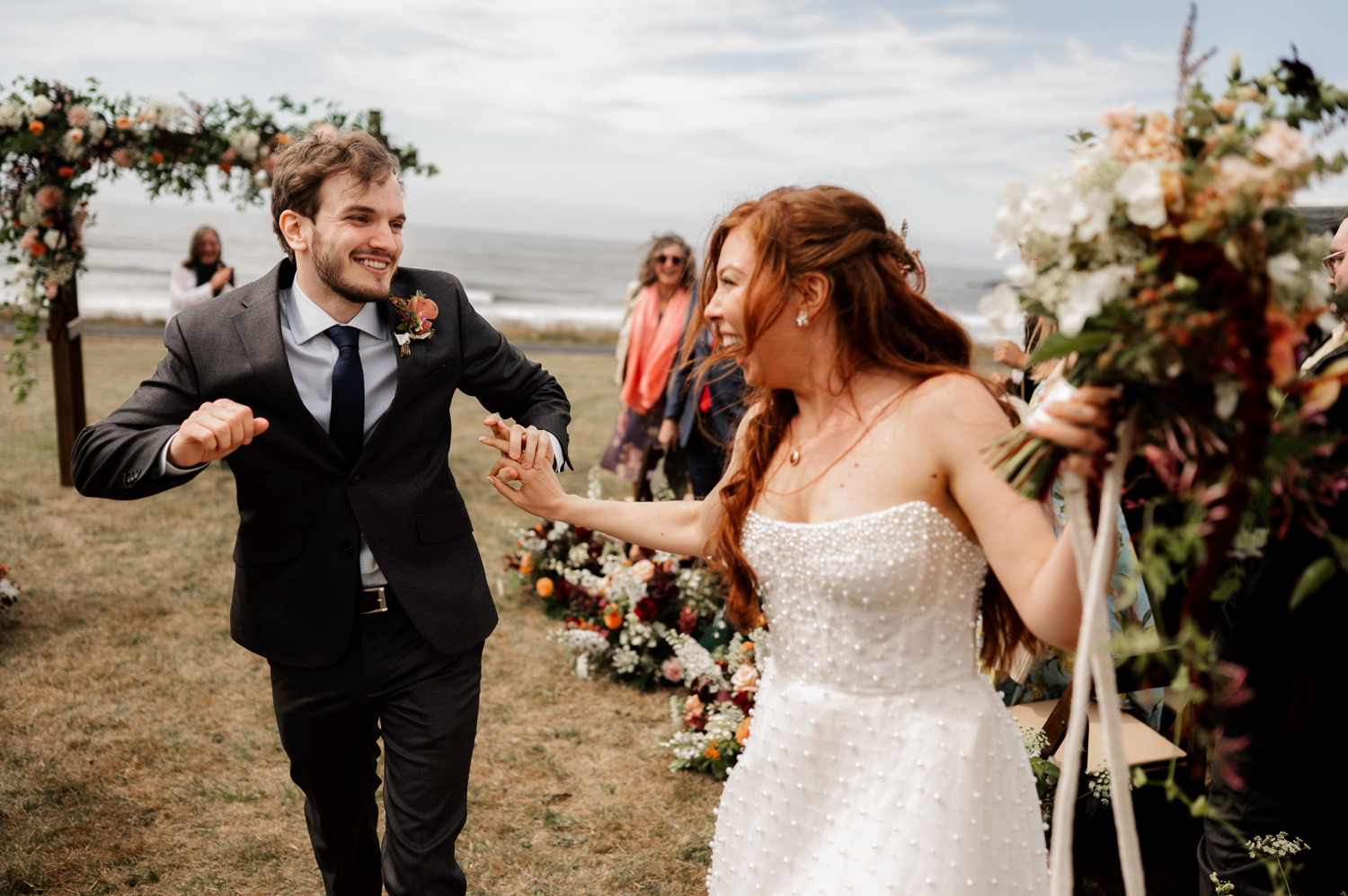 A bride and groom joyfully dance outdoors near a flower-decorated arch with the ocean in the background at the Yachats Inn.
