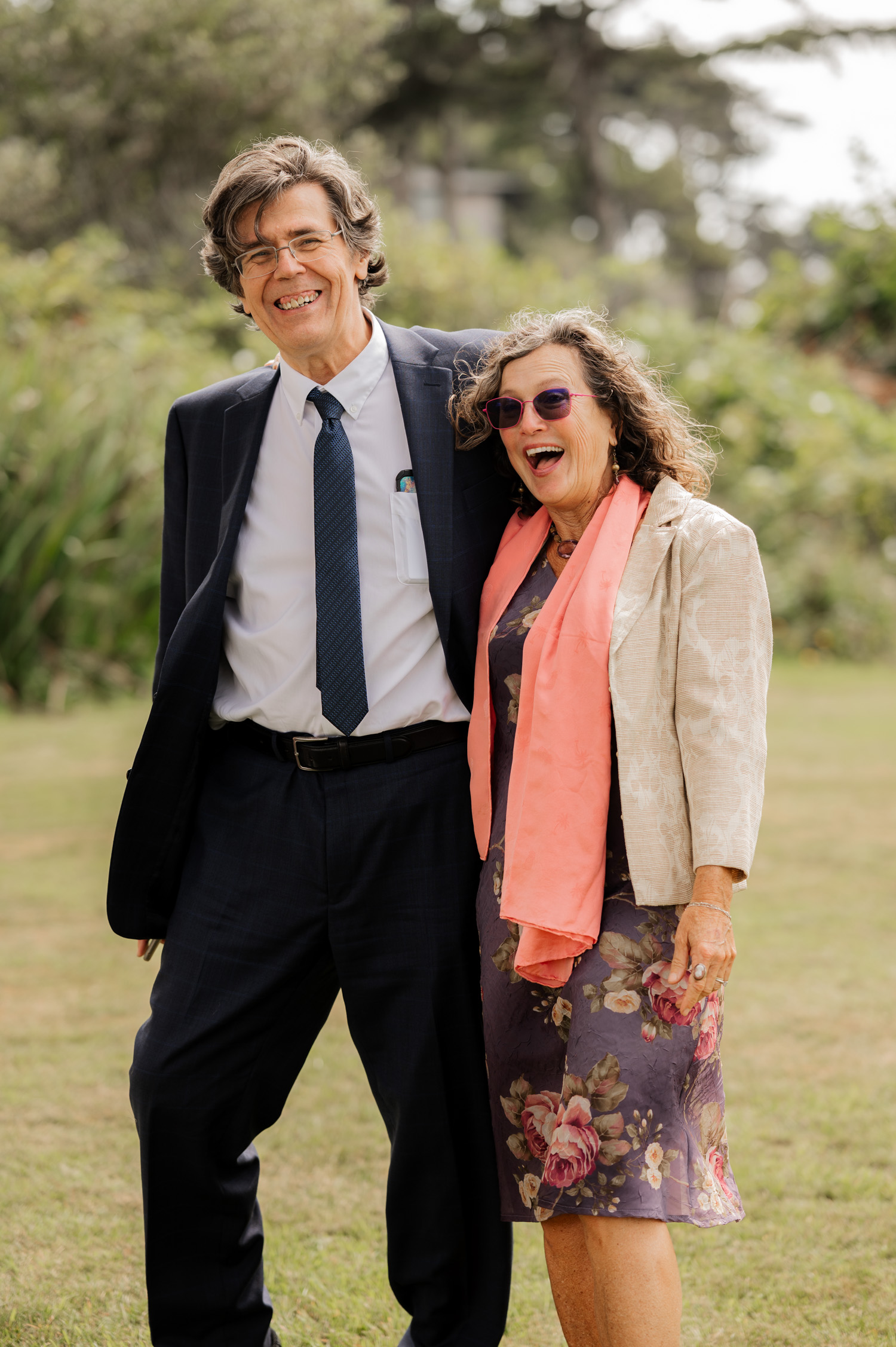 Two people smiling and standing together outdoors at the Yachats Inn, one in a suit and tie, the other in a dress and scarf, taking wedding photos.