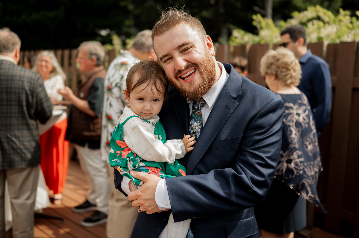 A man in a suit joyfully holds a small child outdoors at a social gathering, with trees and people in the background at the Yachats Inn.