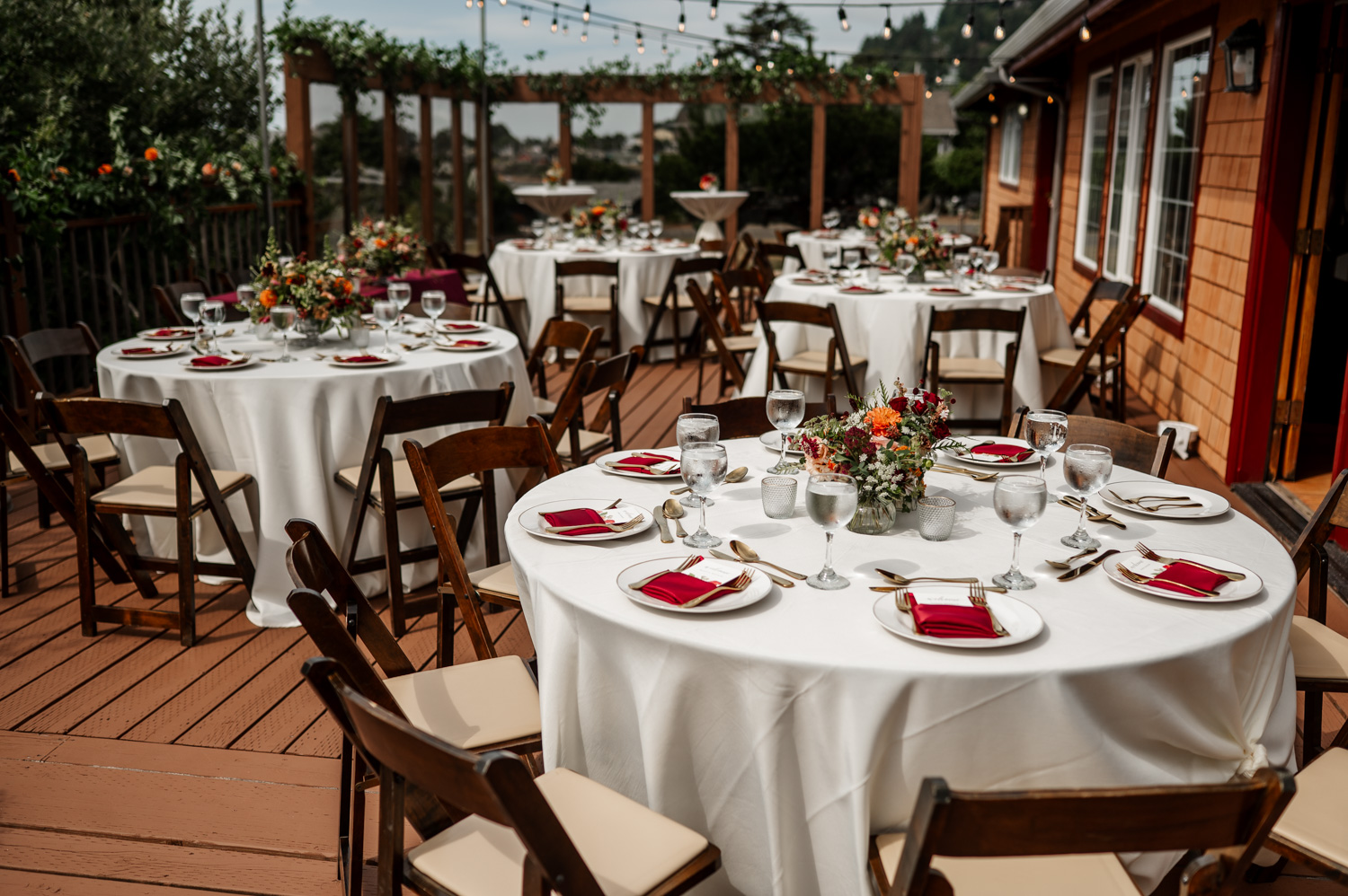 Capture unforgettable wedding photos at the Yachats Inns outdoor dining area. The scene includes round tables with white tablecloths, red napkins, and floral centerpieces under a canopy of string lights.