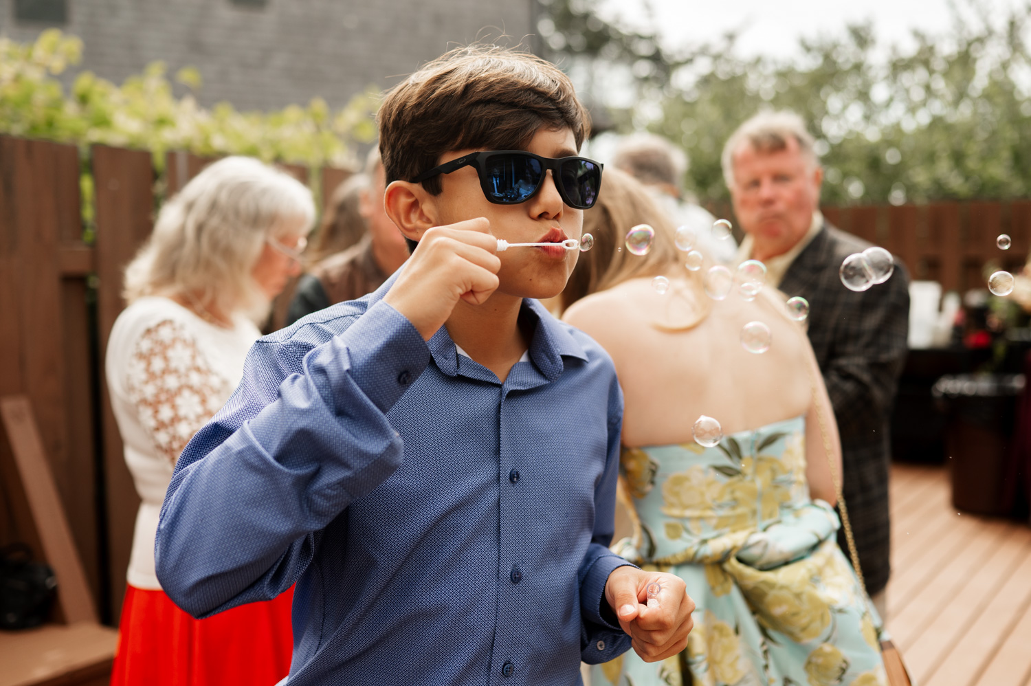 A boy in a blue shirt and sunglasses joyfully blows bubbles outdoors during a wedding at the Yachats Inn, with guests mingling in the background.