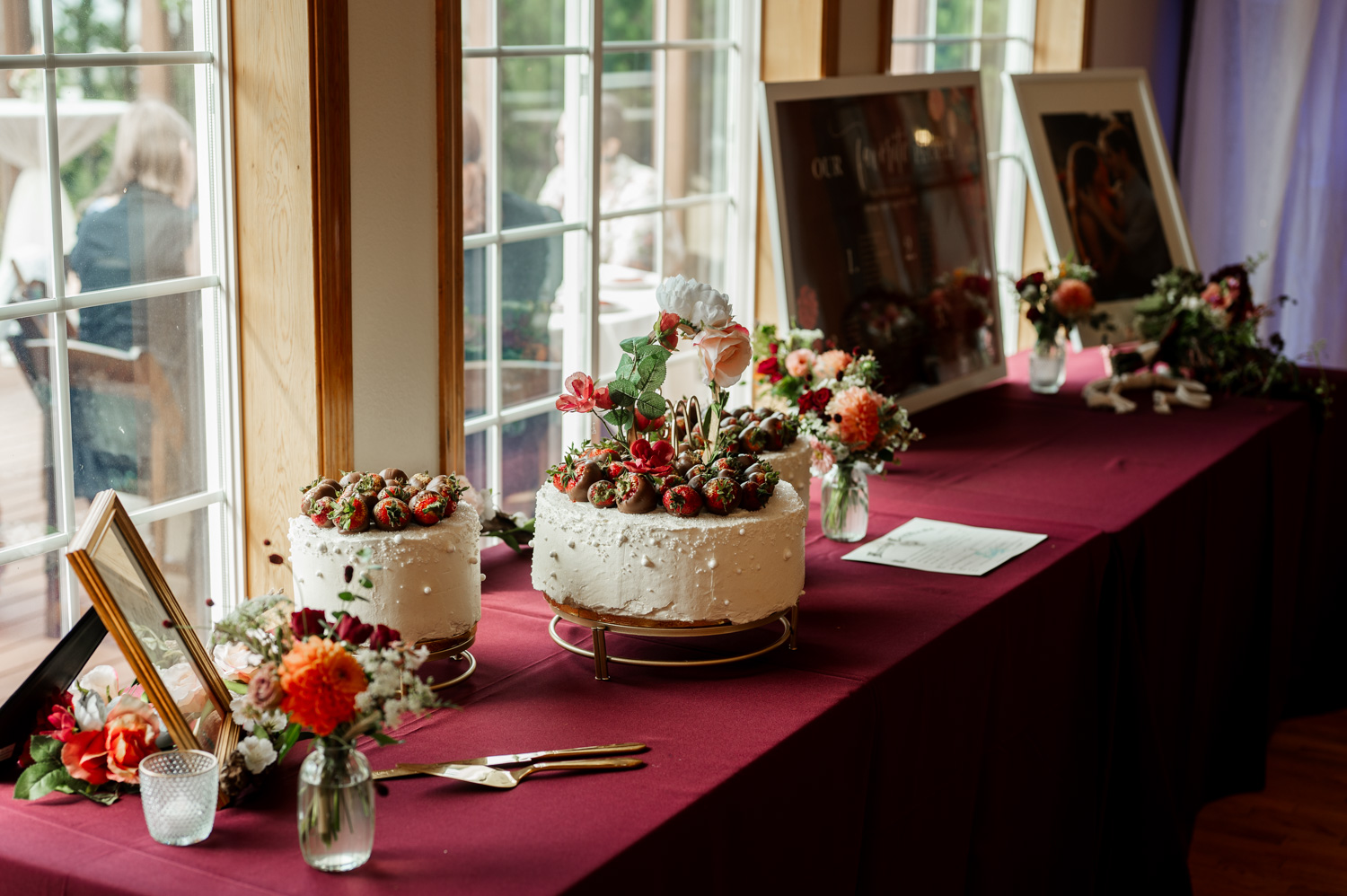 Two decorated cakes are placed on a maroon table, surrounded by flowers and framed wedding photos, next to a window at the Yachats Inn.