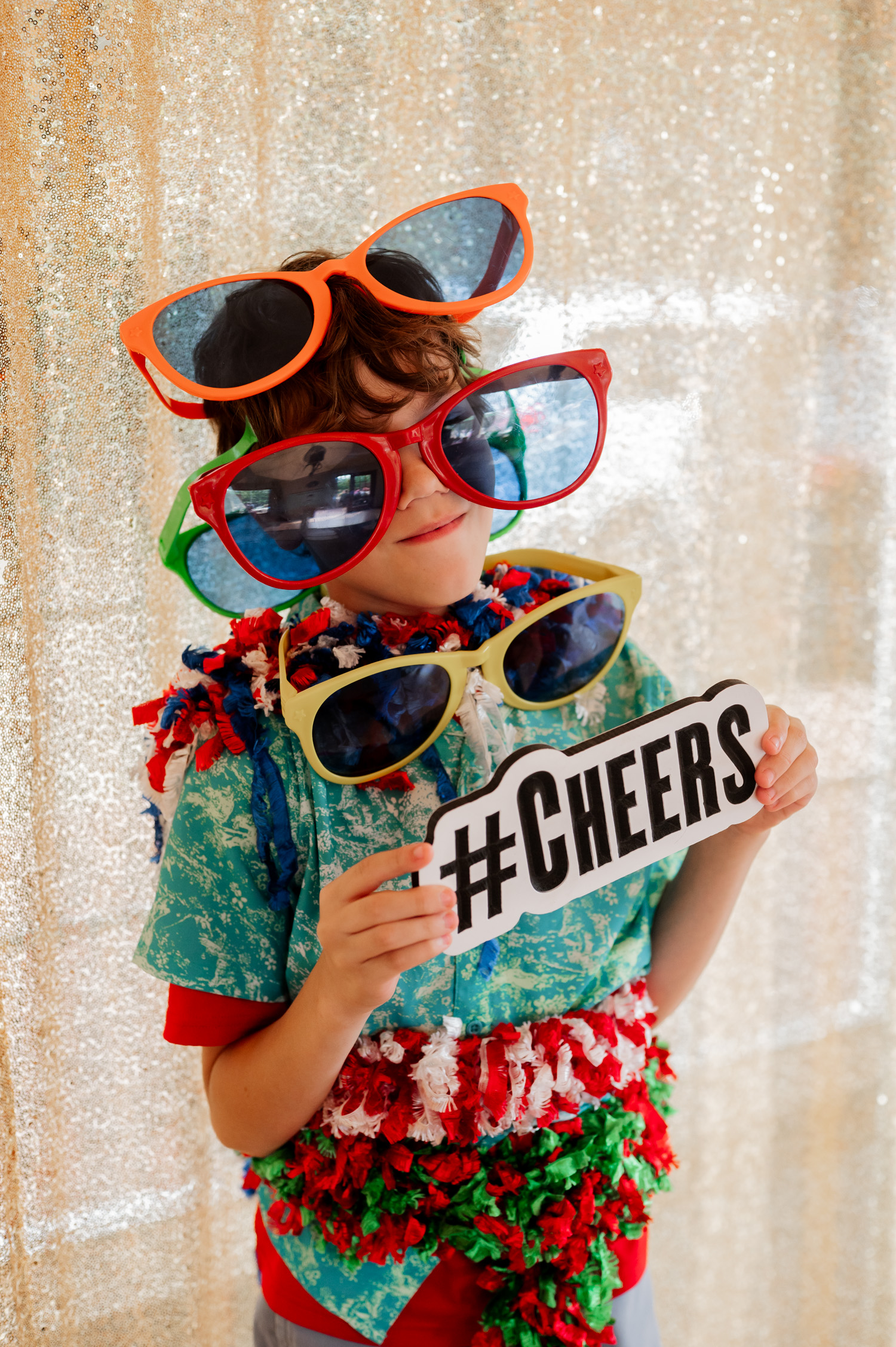 A child wearing oversized sunglasses holds a #CHEERS sign with a sparkly backdrop behind, displaying excitement and readiness for wedding photos at the Yachats Inn.