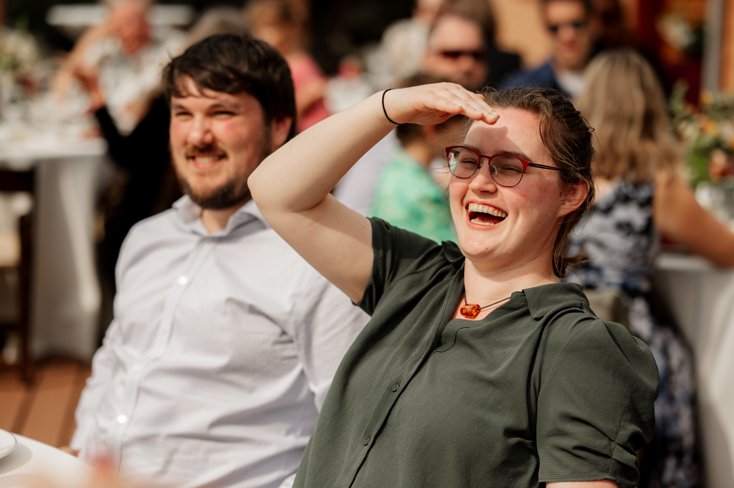 A smiling woman shades her eyes from the sun while sitting next to a man at an outdoor event with white chairs and decorations typical of wedding settings.