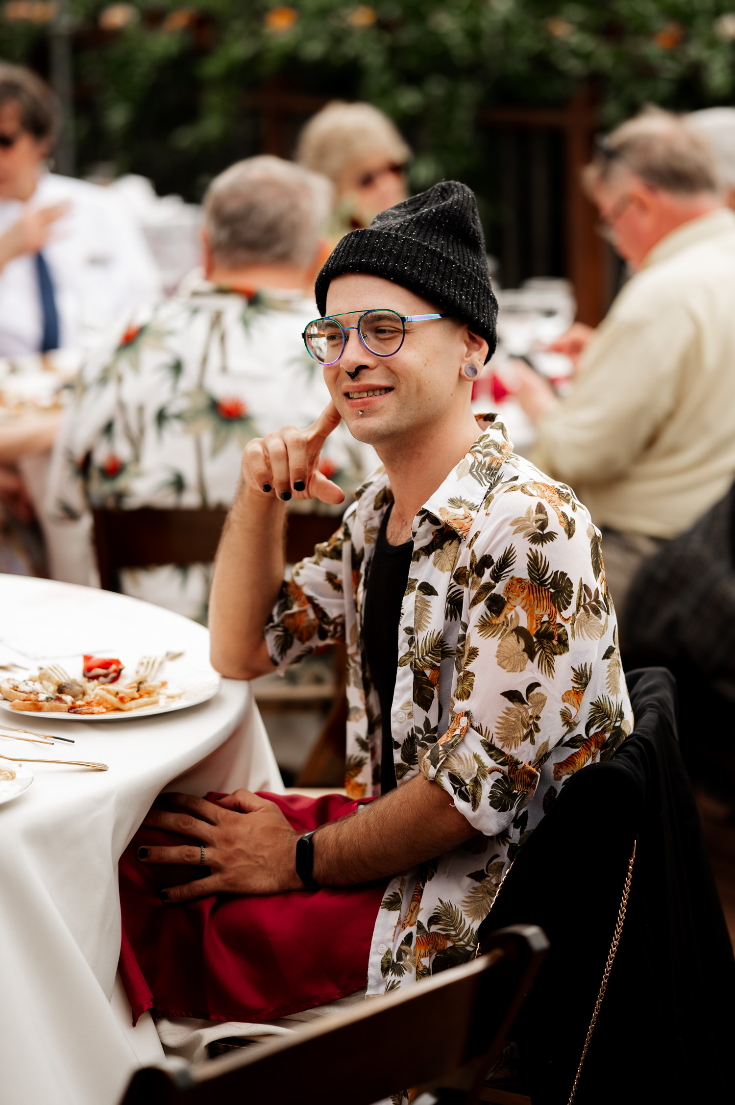 A person in a floral shirt and beanie smiles at an outdoor event, seated at a table with food. The setting appears to be part of wedding photos taken at the Yachats Inn.