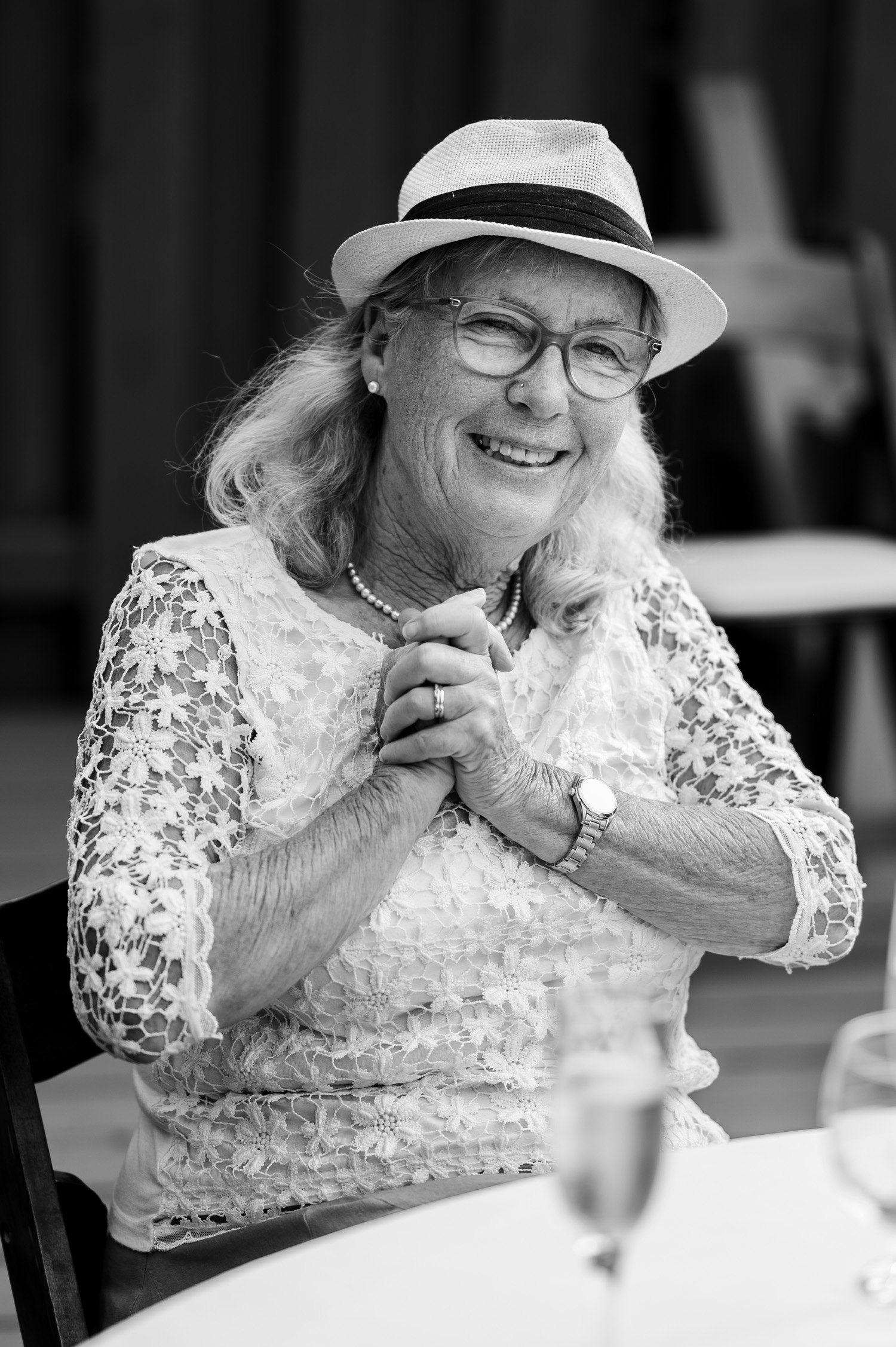 An elderly woman in a hat and lace blouse smiles at a table with a glass, hands clasped. She looks at wedding photos, possibly at the Yachats Inn. The image is in black and white.