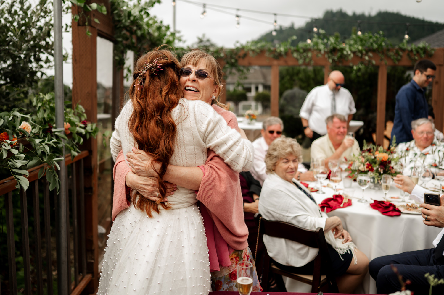 A bride joyfully embraces an older woman at the outdoor reception of the Yachats Inn, surrounded by seated guests and vibrant greenery in the background.