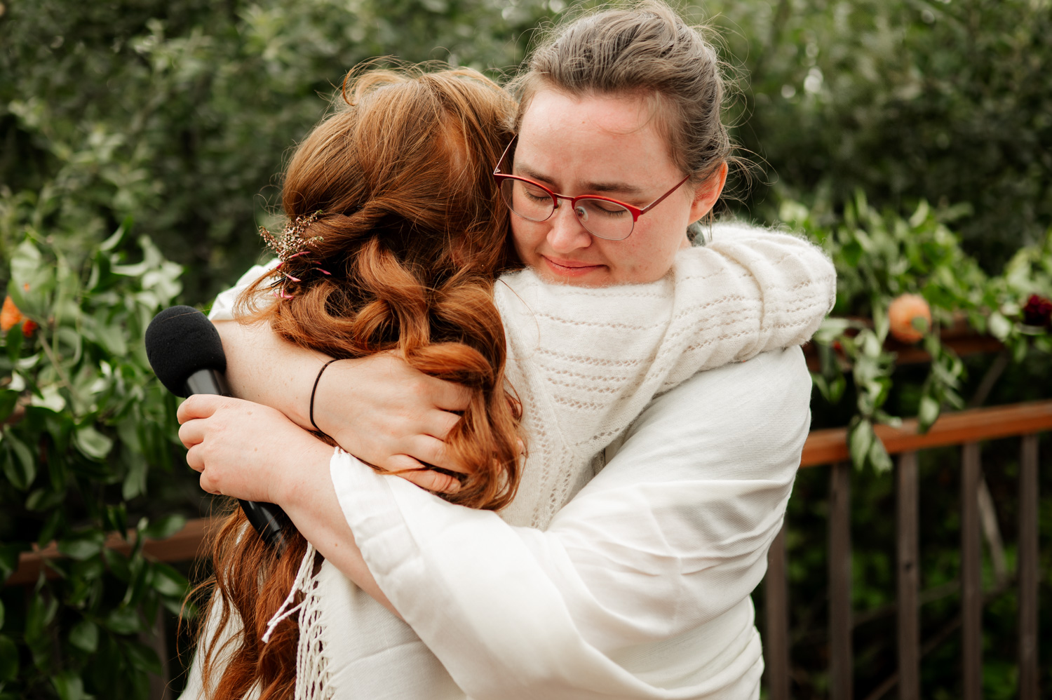 Amid lush greenery, two people share a warm embrace outdoors, with one person holding a microphone. They appear to be in a heartfelt moment, reminiscent of wedding photos.