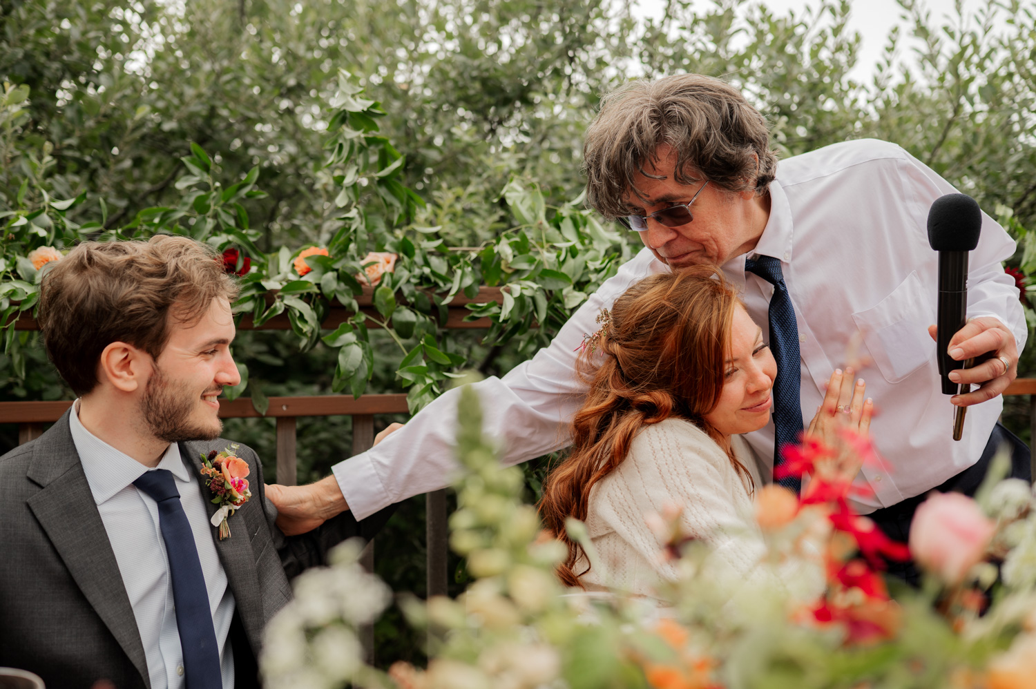 A man with a microphone hugs a seated woman as another man in a suit smiles at them, surrounded by flowers at the Yachats Inn wedding.