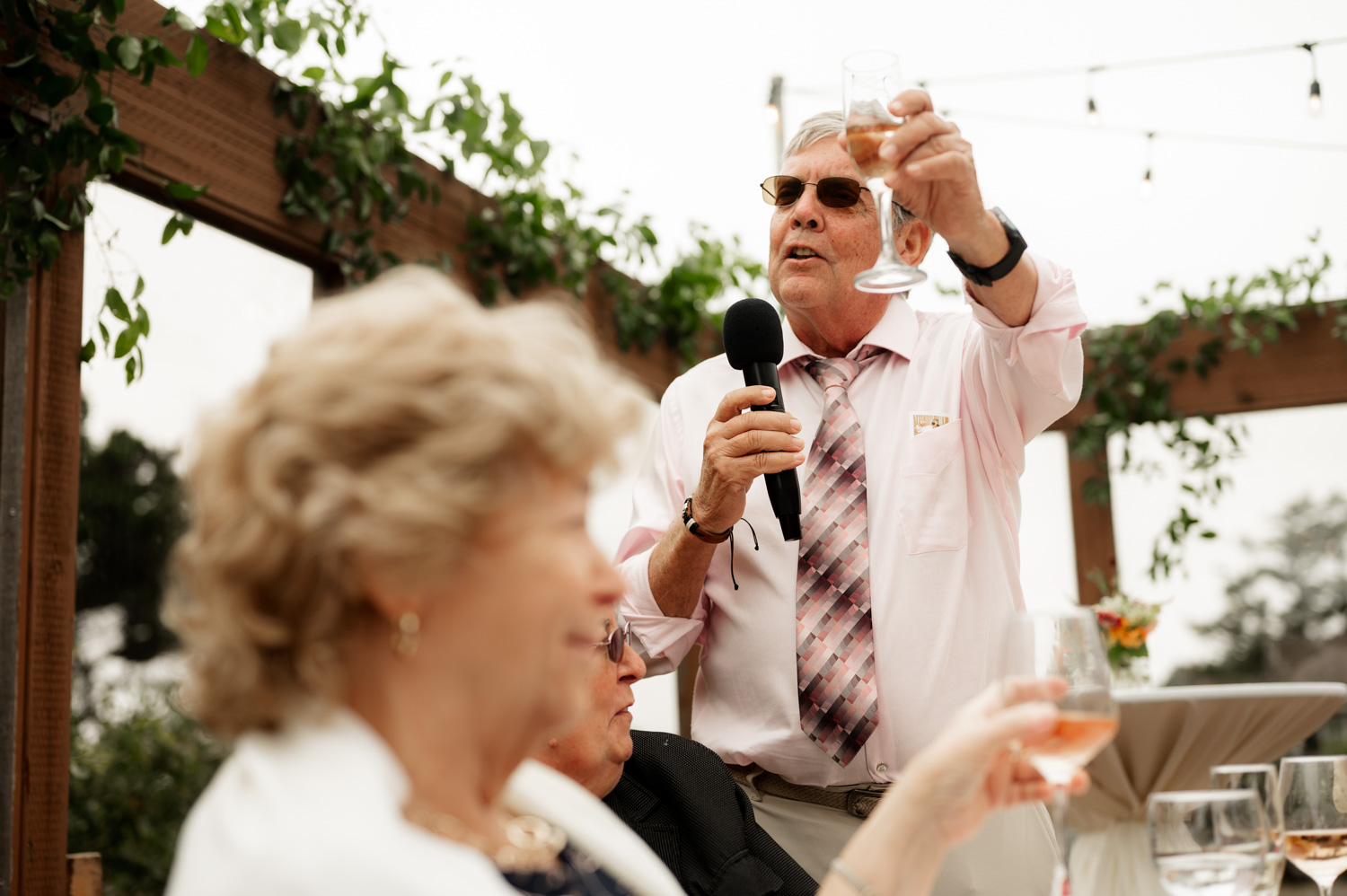An elderly man holds a microphone and gives a toast as guests raise their glasses at the Yachats Inn, with a scenic backdrop visible at this outdoor wedding event.