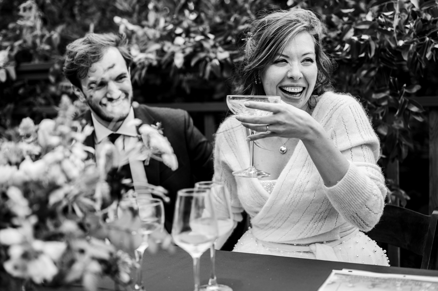 A bride and groom laughing as the grooms face is covered in cake. They are surrounded by flowers and drink glasses at their wedding at the Yachats Inn.