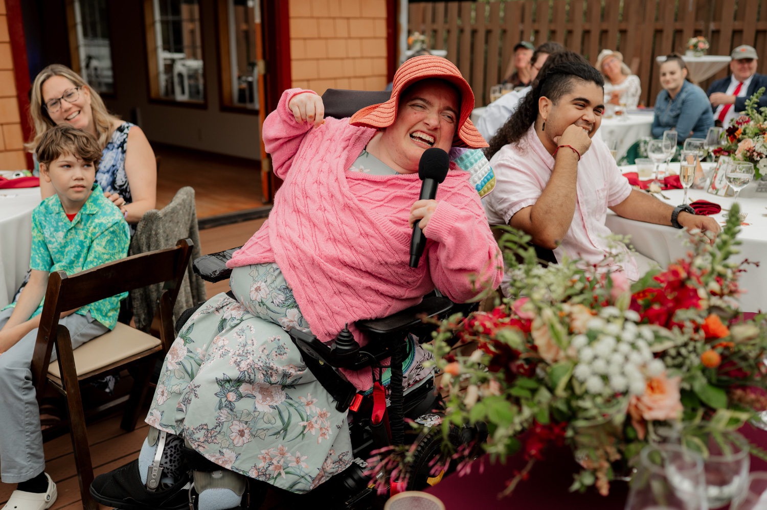 A person in a wheelchair joyfully speaks into a microphone at an outdoor event, surrounded by guests and flowers at the Yachats Inn.