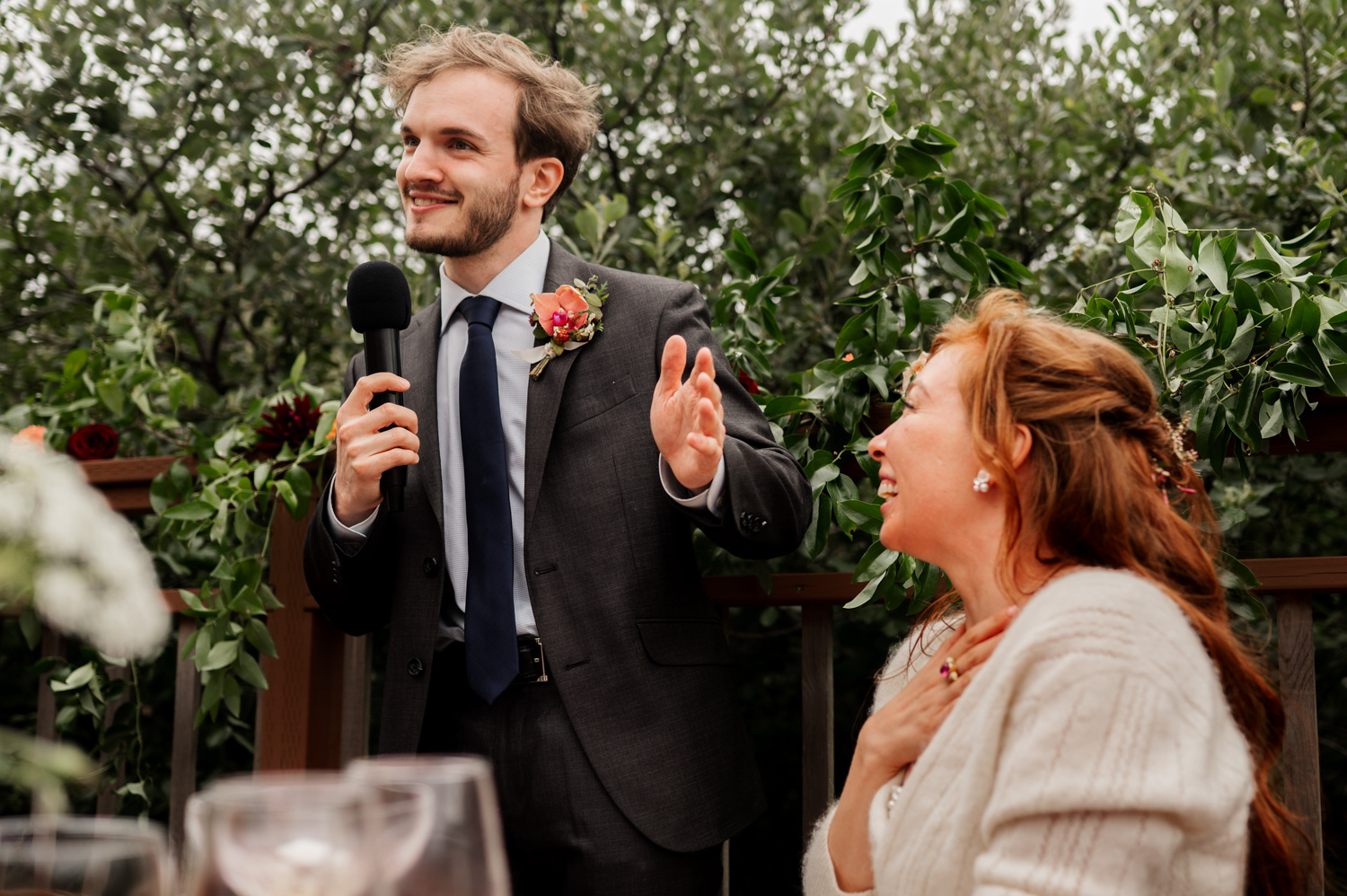 A man in a suit holds a microphone and gives a heartfelt speech next to a smiling woman, surrounded by the lush greenery at the Yachats Inn, during memorable wedding moments.