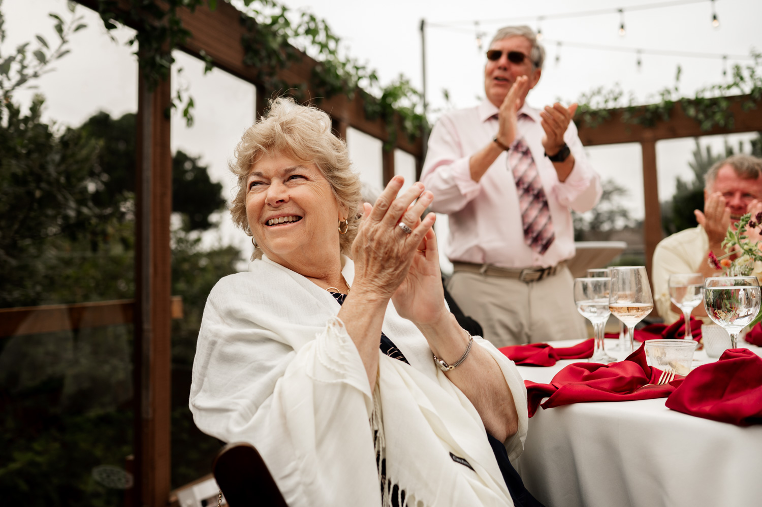 A woman smiling and clapping at an outdoor event, with joyful guests and elegantly arranged tables in the background.
