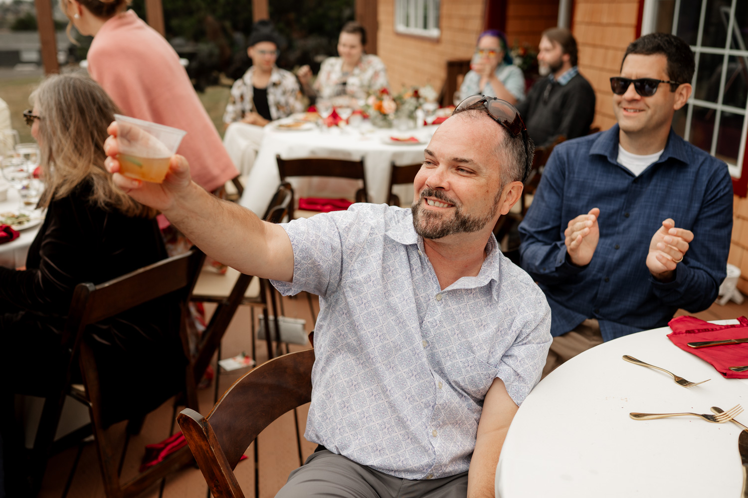 A man smiles and raises a drink at an outdoor event, with guests seated at elegantly decorated tables in the background.