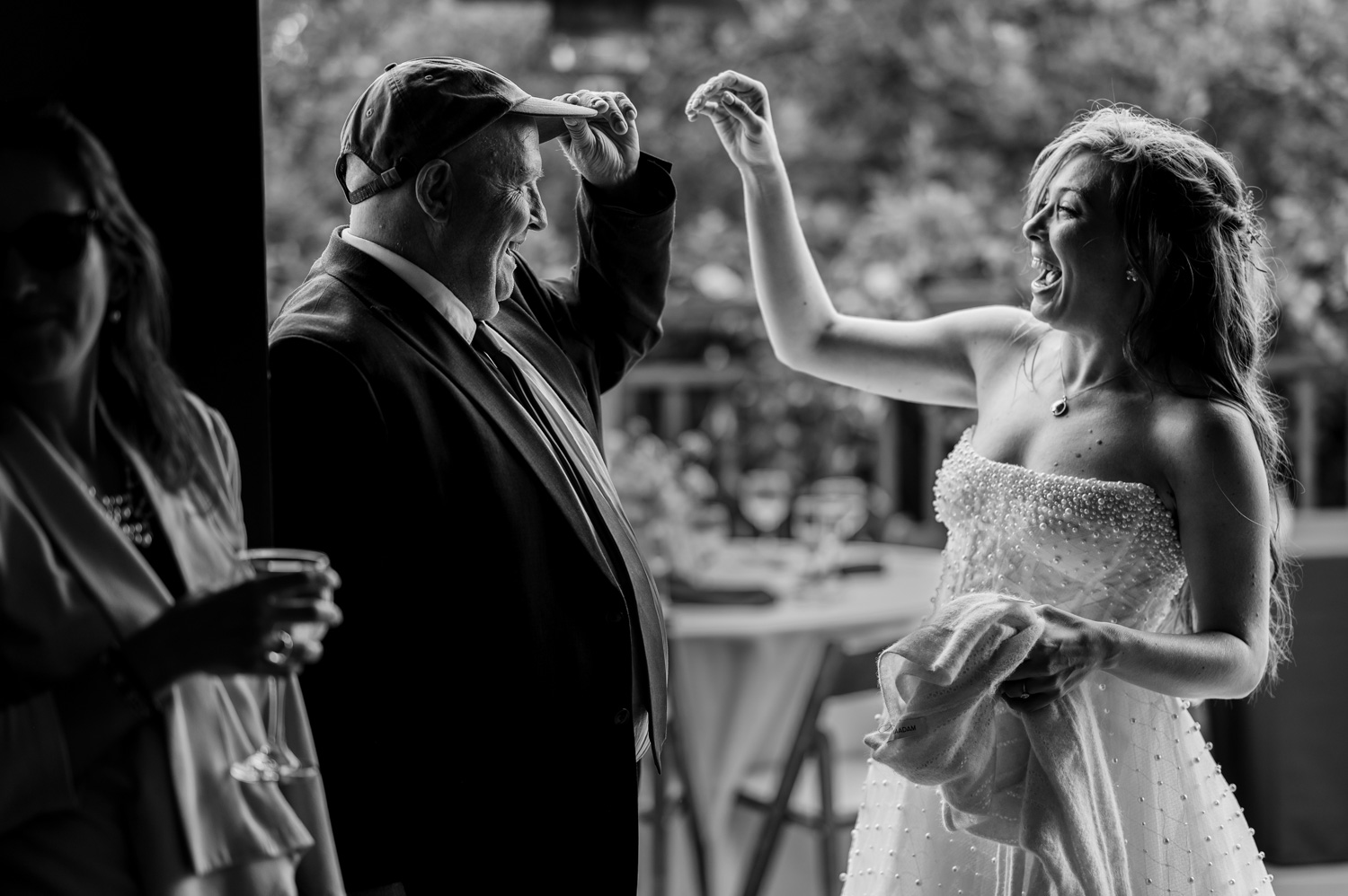 A bride in a white gown smiles brightly as she interacts with a man wearing a cap, surrounded by lush indoor greenery at the Yachats Inn.