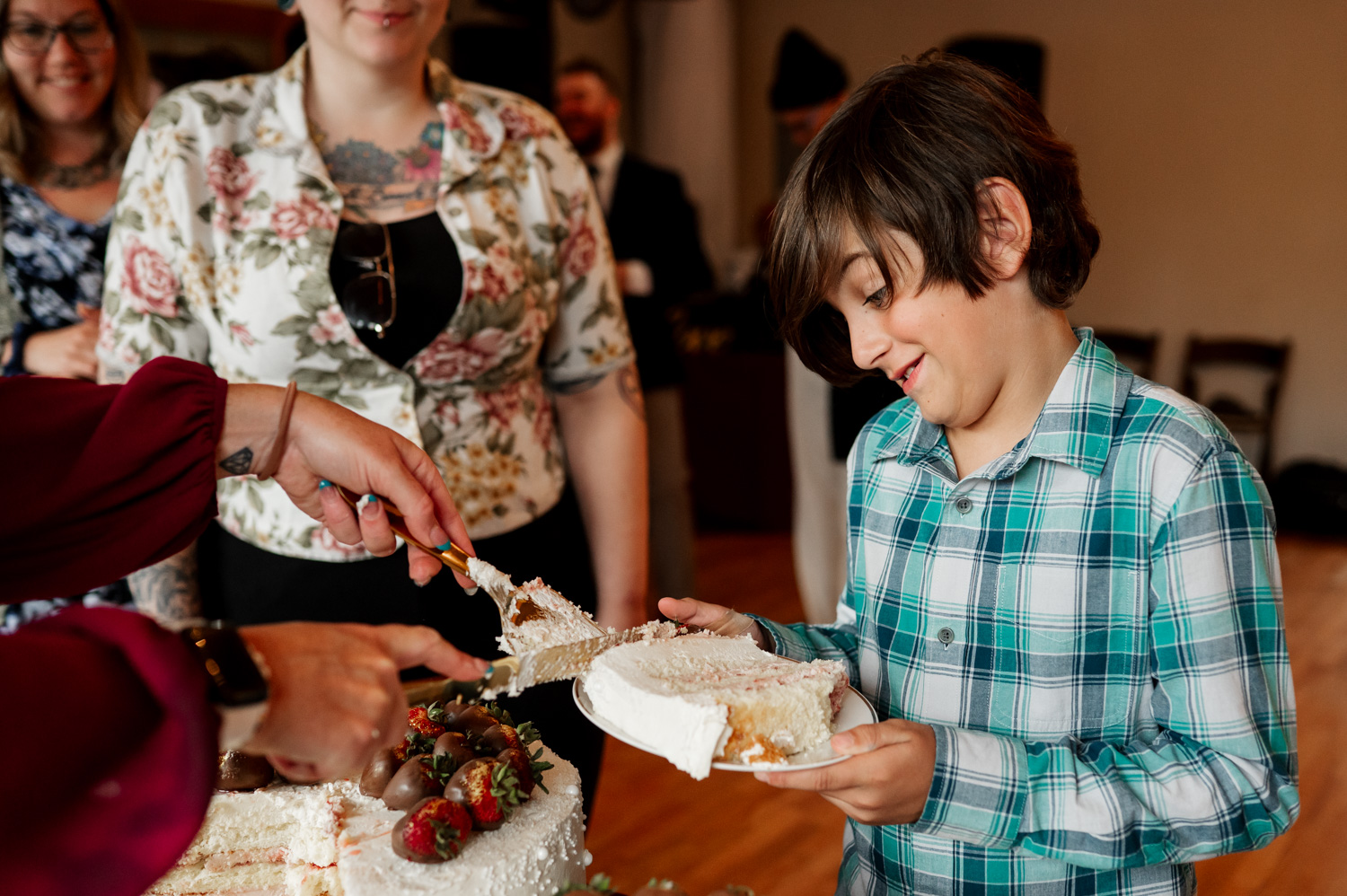 A joyful celebration with a child in a plaid shirt eagerly holding a plate for cake being served; people in the background are smiling and laughing.