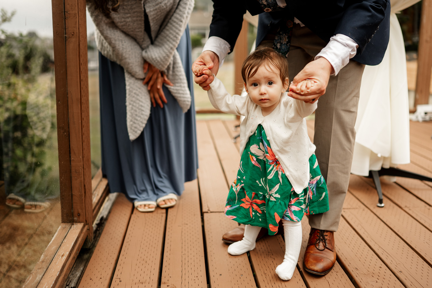 A toddler in a floral dress is assisted in walking by a man as a woman observes nearby on a wooden deck during wedding photos at the Yachats Inn.