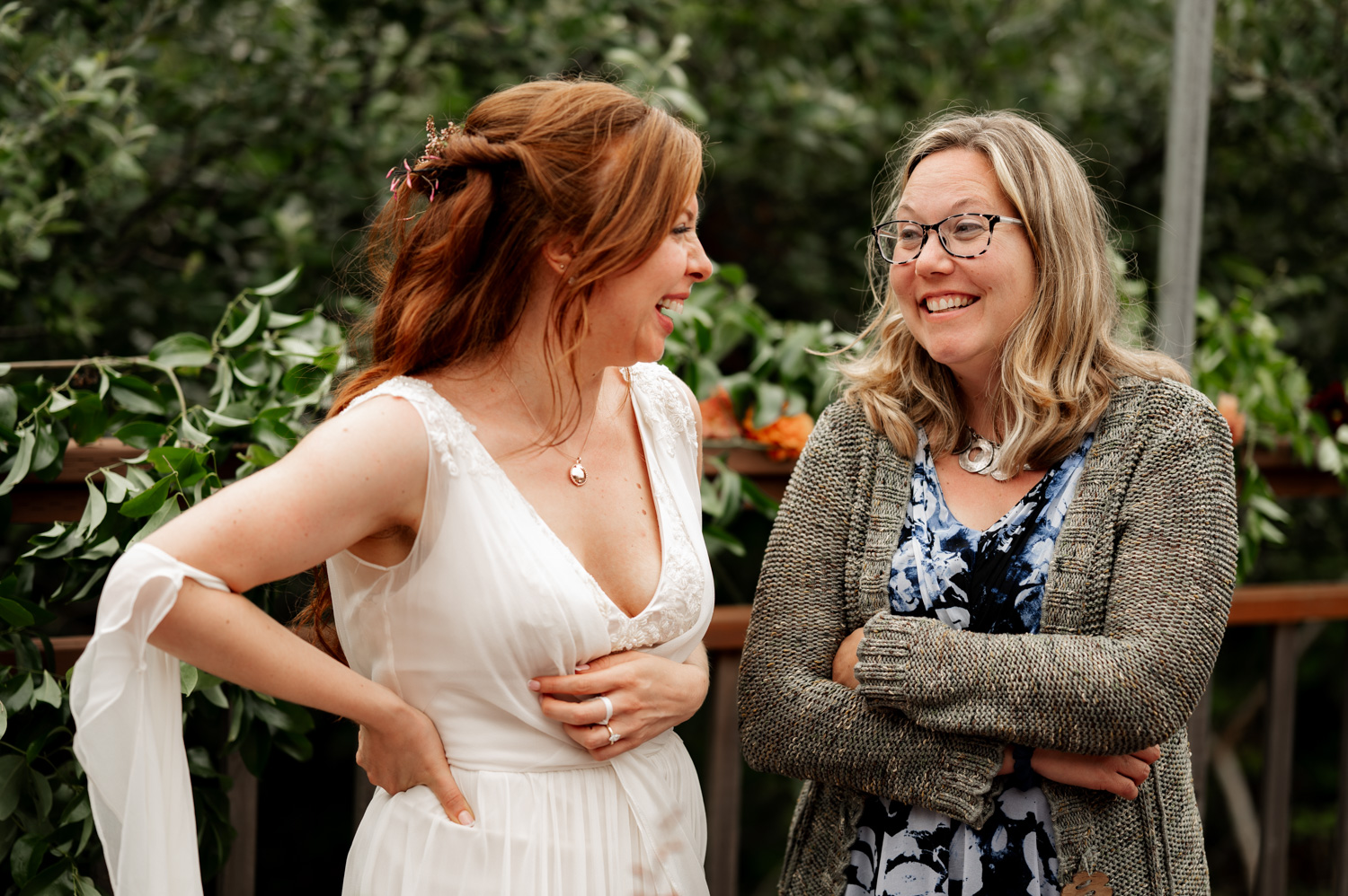 Two women laughing together outdoors, one wearing a white dress and the other in a patterned dress with a cardigan, standing near the Yachats Inn.