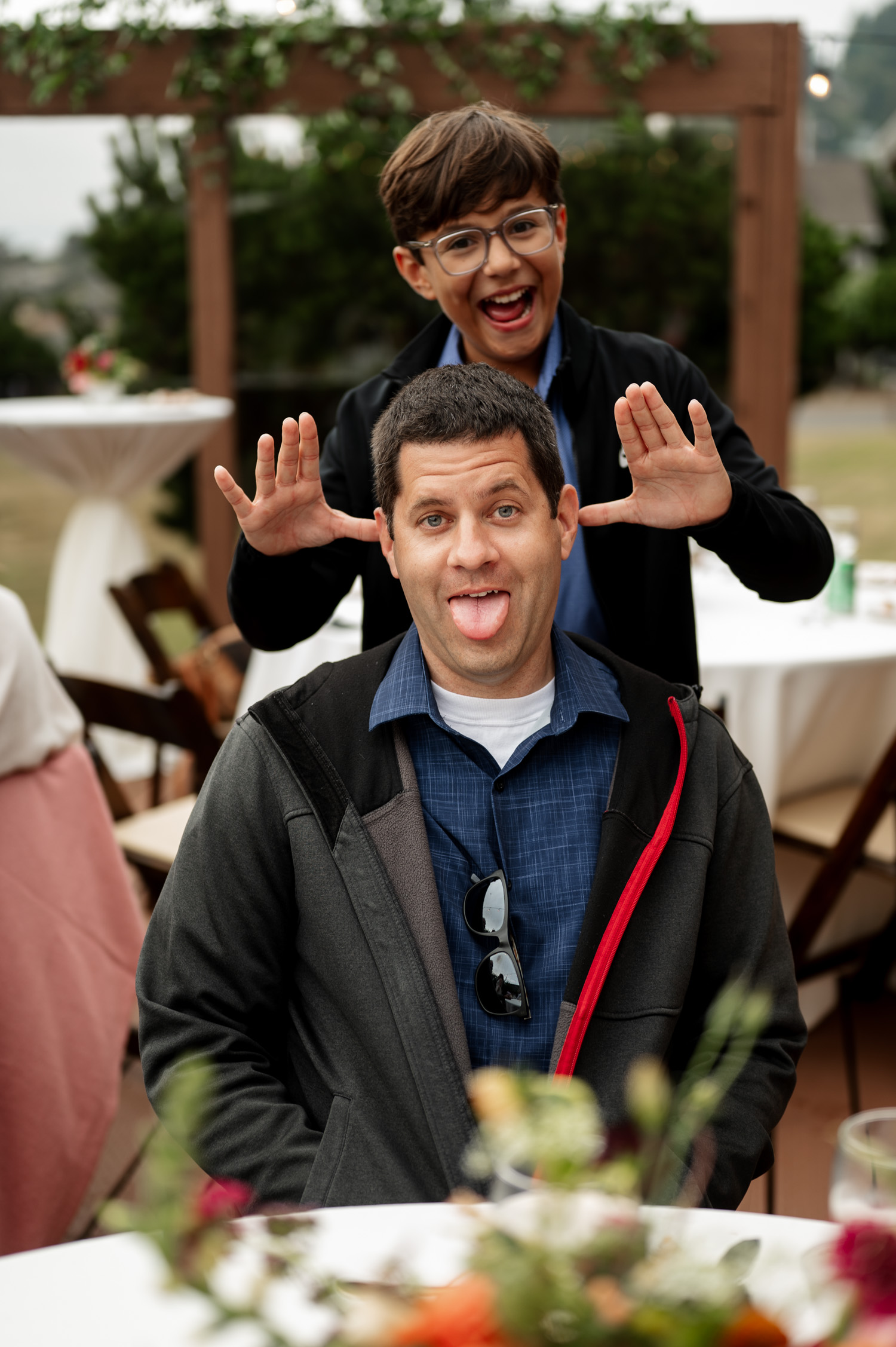 Two people at an outdoor event, one smiling playfully behind the other who is sticking out their tongue, with a background displaying the setting of Yachats Inn during a wedding celebration.