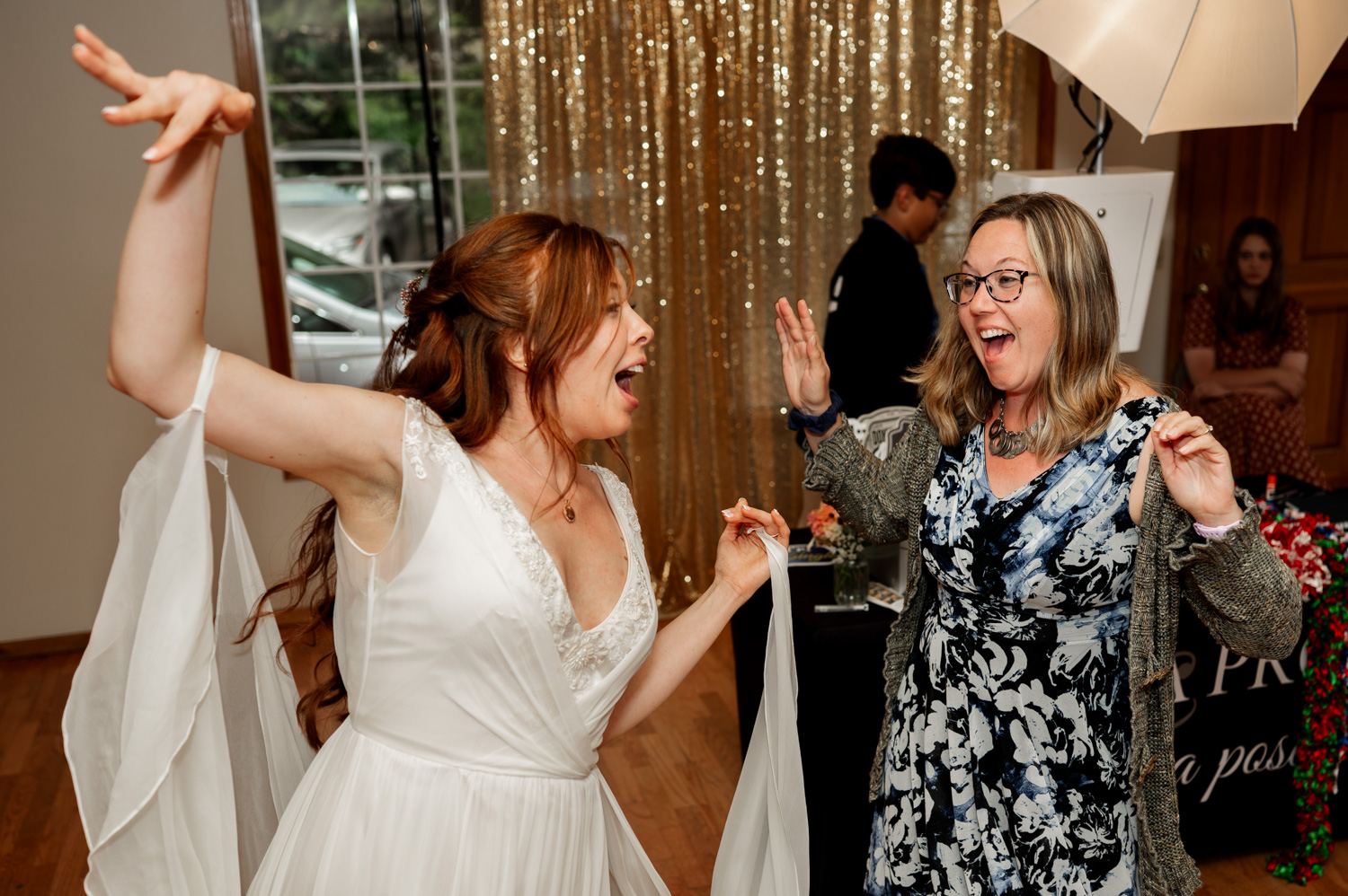 Two women dance joyfully, one in a white dress and the other in a floral outfit, against a gold backdrop at the Yachats Inn.