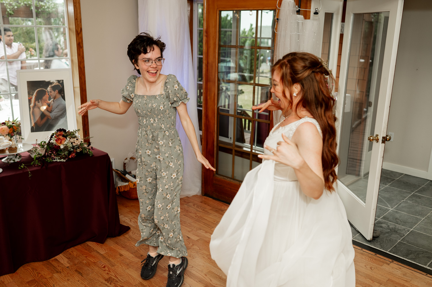 Two women dancing indoors, one wearing a floral dress and the other in a white gown, with wedding photos displayed on the walls at the Yachats Inn.