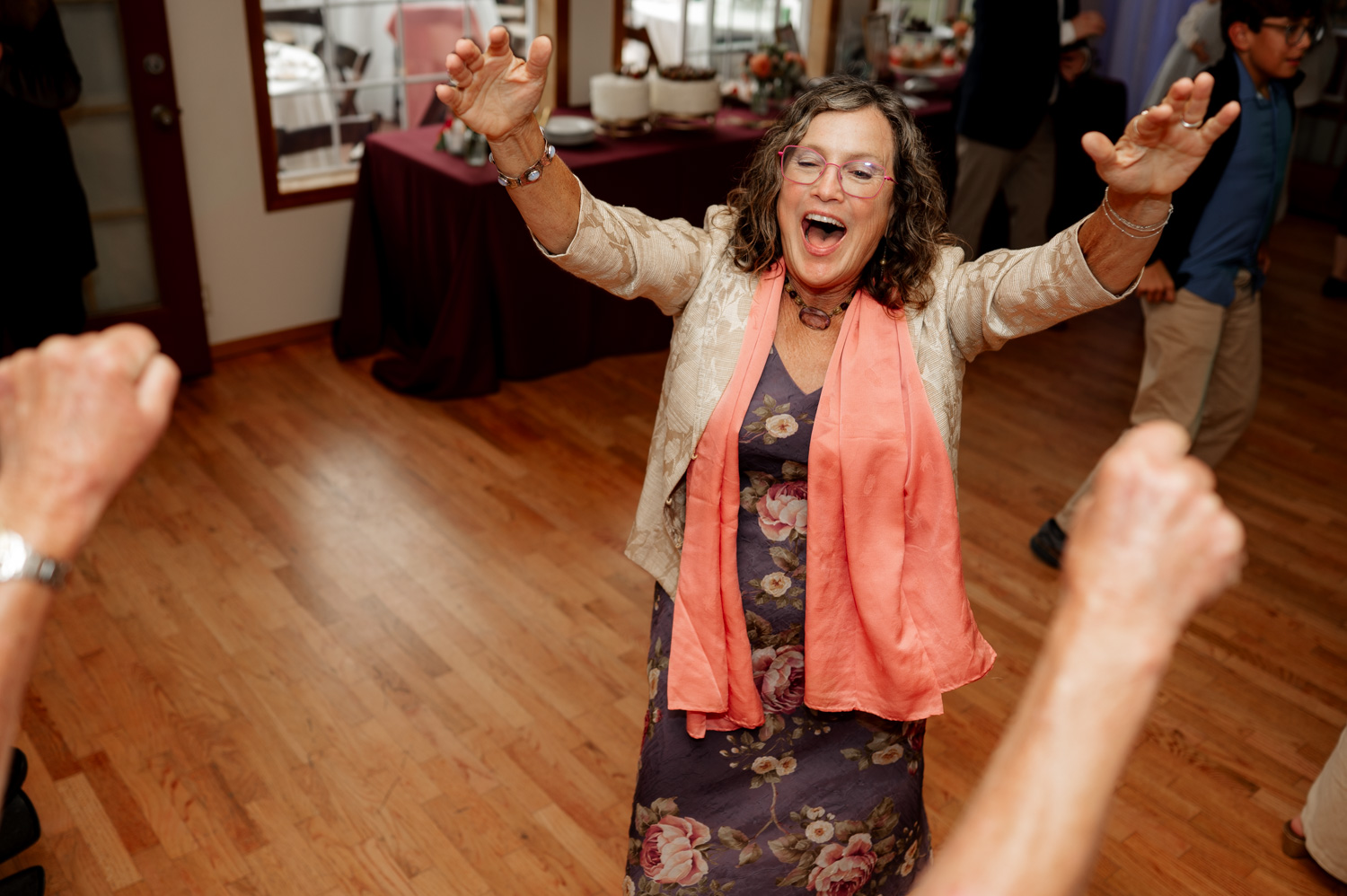 A joyful woman with raised arms and a smile, wearing a floral dress and peach scarf, dances on the wooden floor at the Yachats Inn during a wedding celebration.