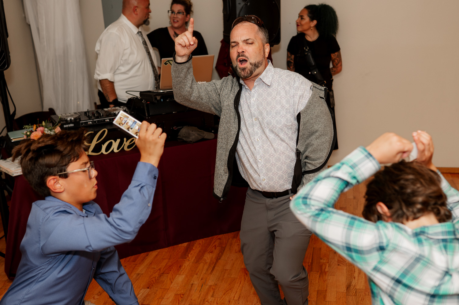 At the Yachats Inn, a man energetically dances at an event while others watch. A child with glasses takes a photo of the moment with a smartphone, blending into the wedding photos.