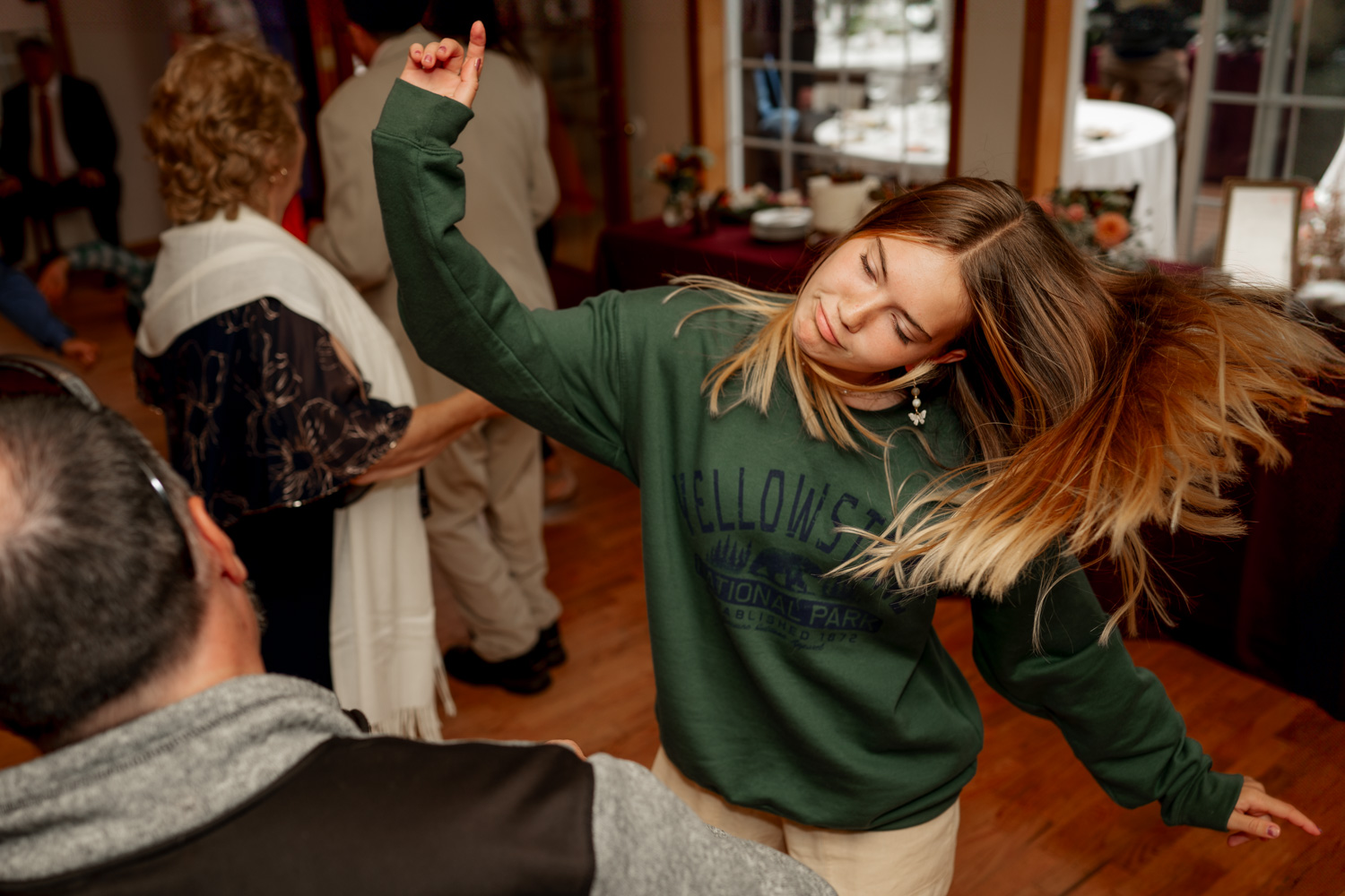 A woman in a green sweatshirt dances energetically indoors, her hair swaying, with guests from the wedding at Yachats Inn cheering in the background.
