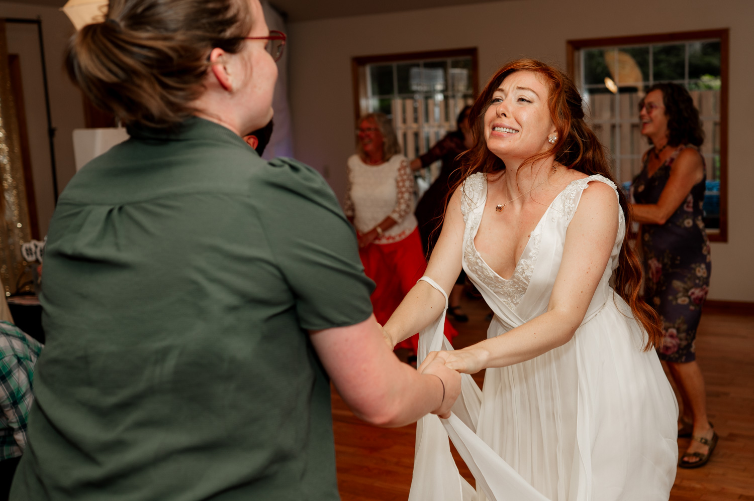 The bride joyfully dances in her white dress, twirling next to a person in a green shirt. In the background, guests mingle happily at the wedding celebration held at the Yachats Inn.