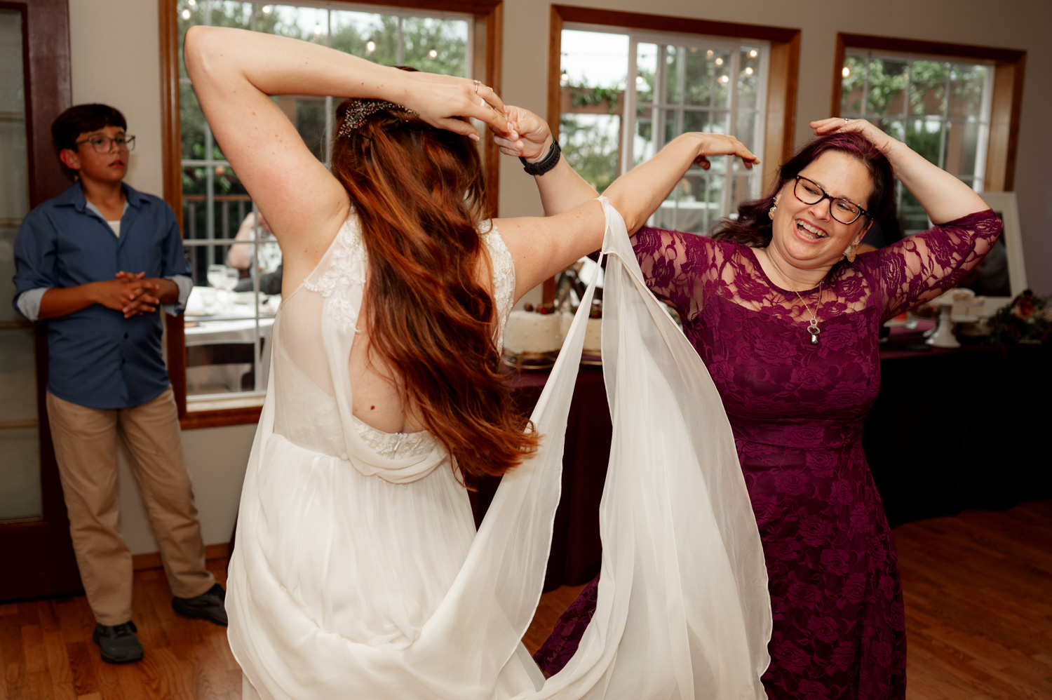 Two women dance joyfully on wooden floors near large windows, their laughter evident, while a man watches from the background with admiration.