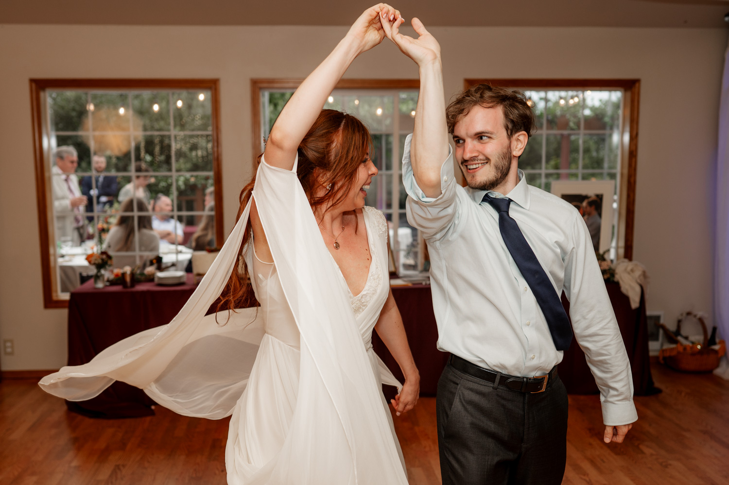 A couple dances joyfully indoors, capturing a moment of pure bliss. The woman twirls in a white dress, while the man complements her with a shirt and tie, evoking enchanting wedding photos at the Yachats Inn.