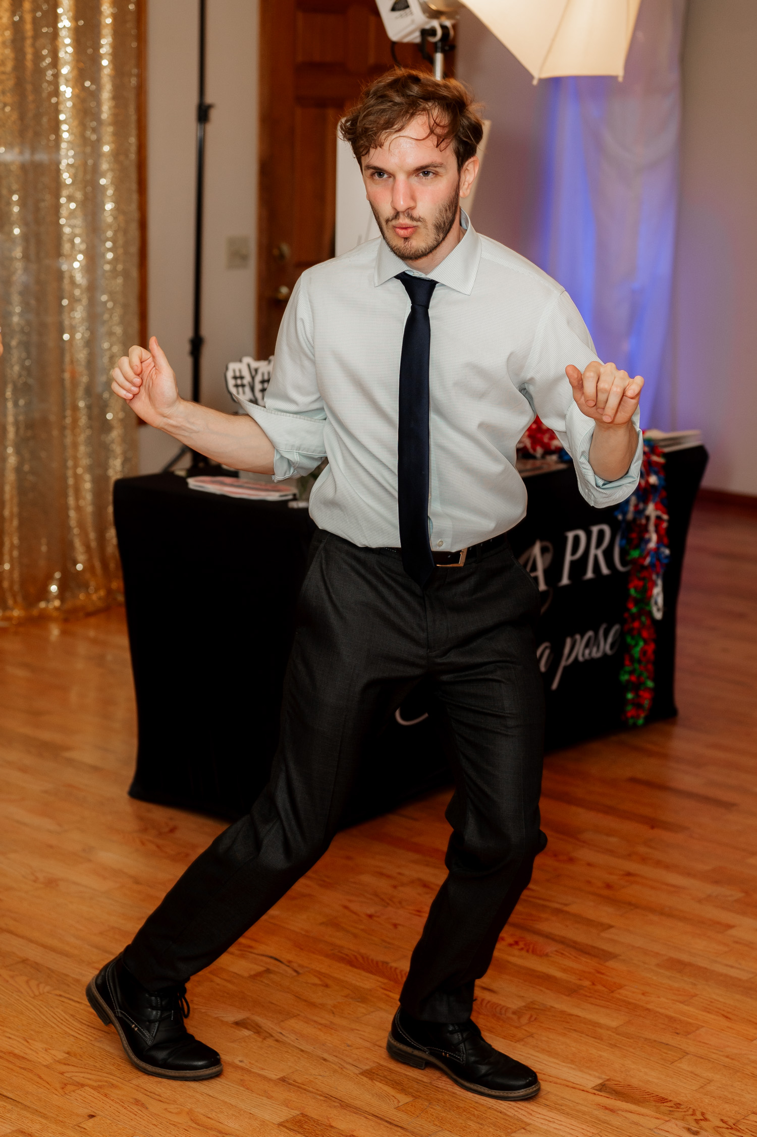 A man in a shirt and tie dancing energetically on a wooden floor in a beautifully decorated room, with people around him laughing and smiling at the Yachats Inn during a wedding.
