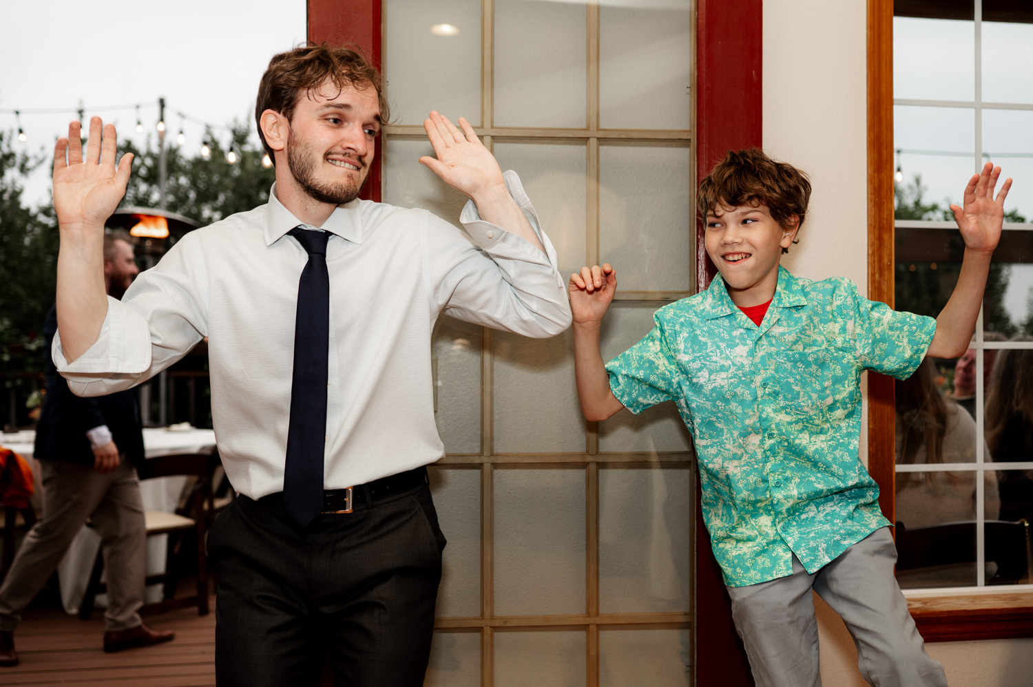 A man and boy in vibrant outfits laughing and posing with raised hands indoors during wedding photos at the Yachats Inn.