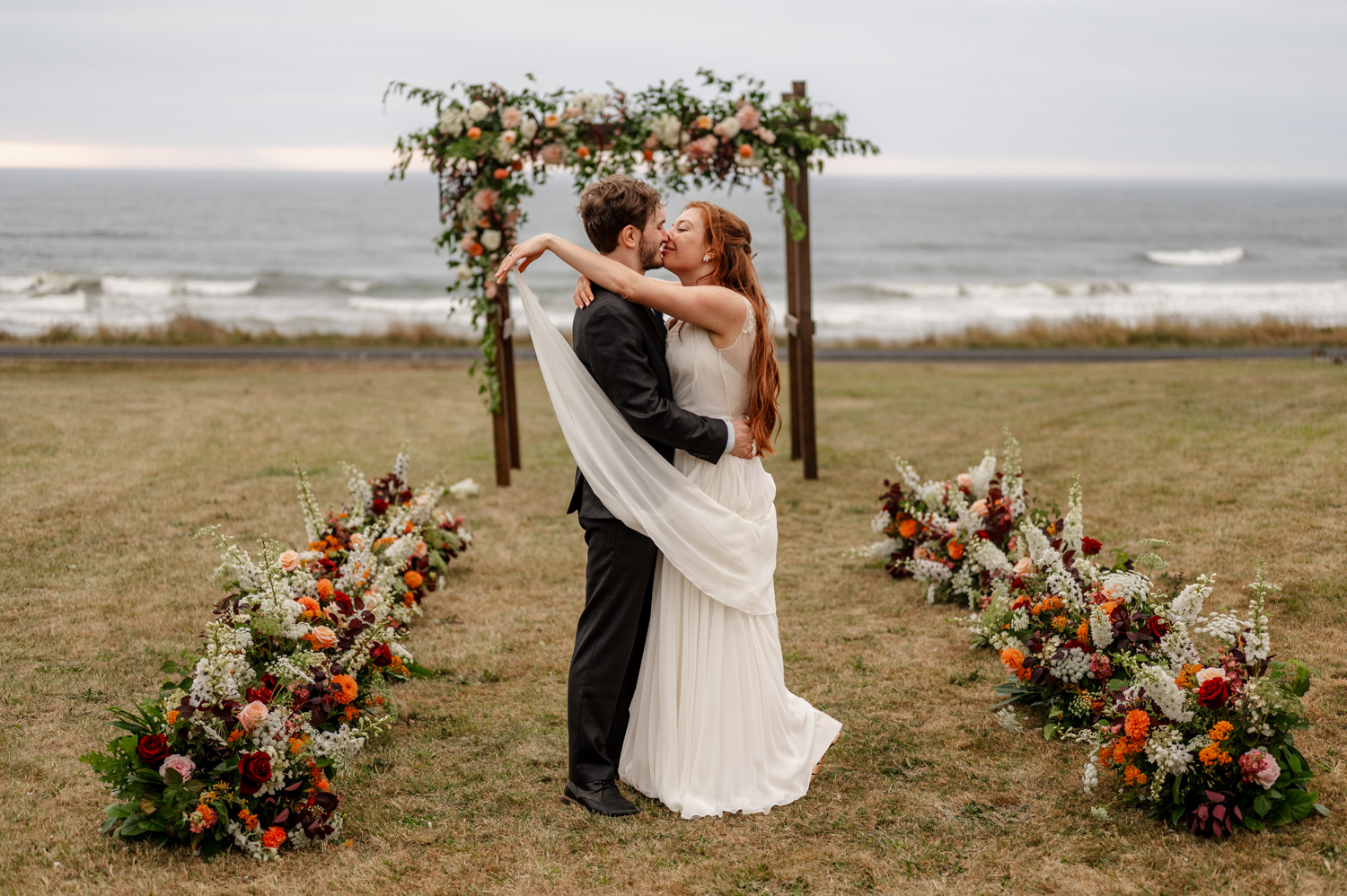 A couple embraces at their oceanfront wedding by the Yachats Inn, with stunning floral arrangements and a charming wooden arch in the background.