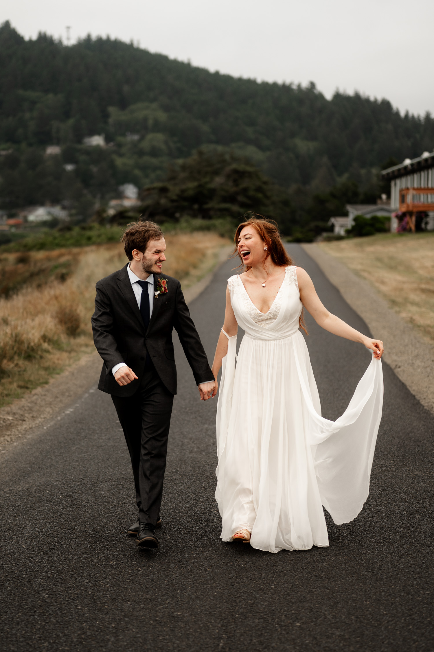 Bride and groom walking hand-in-hand on a road with green hills in the background at Yachats Inn.