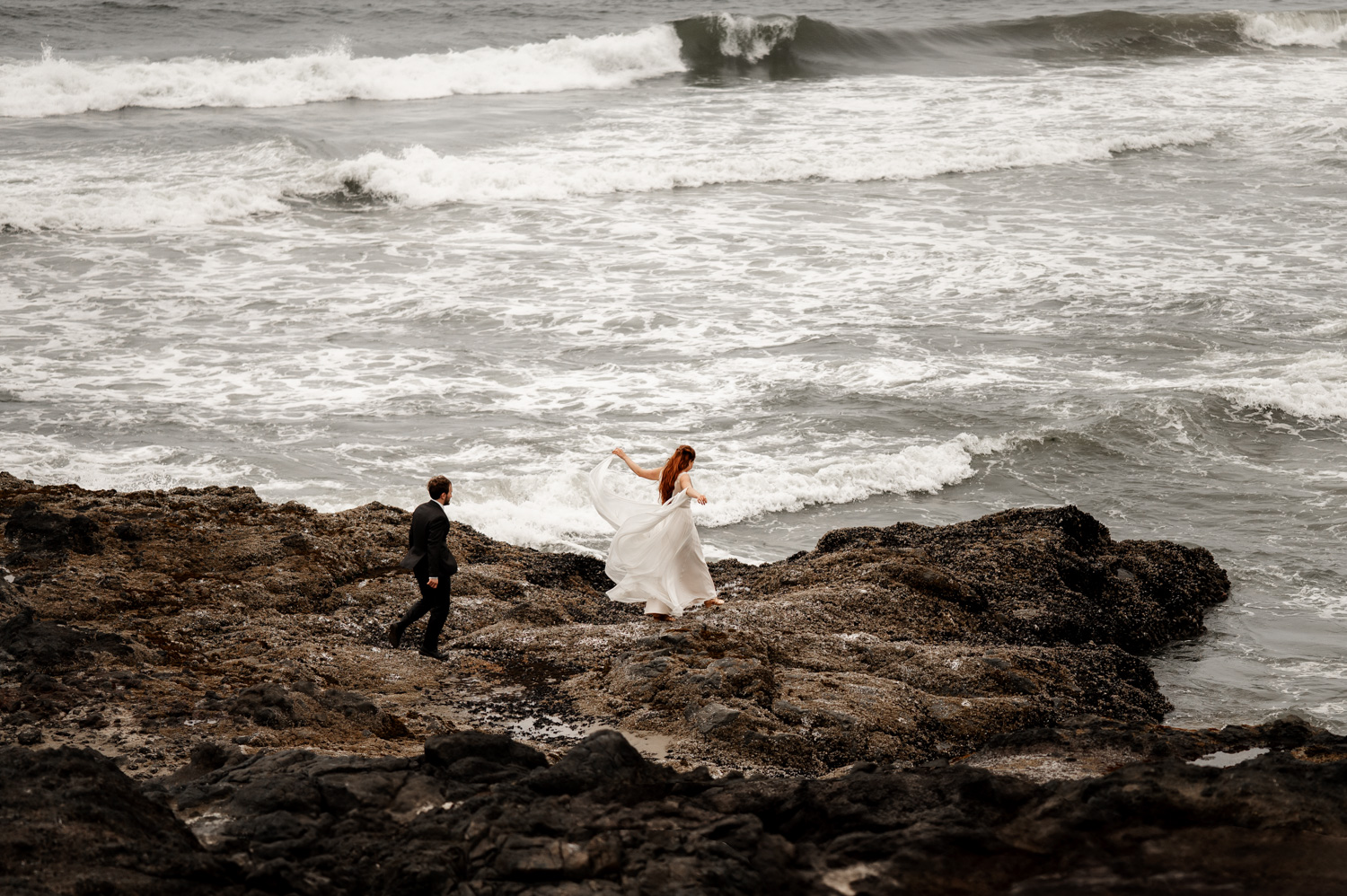A bride and groom walk over rocky terrain by the ocean, with the bride holding her dress, during a wedding photo shoot at Yachats Inn.