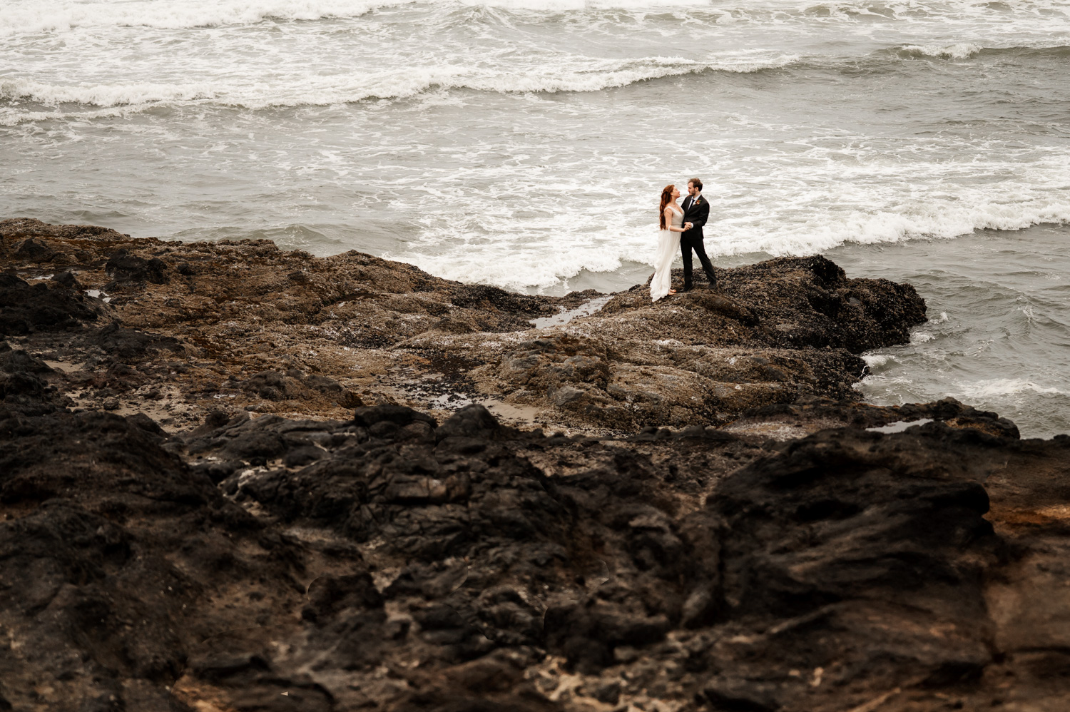 A couple embraces on a rocky shore with waves crashing behind them, capturing a tender moment that resembles wedding photos at the Yachats Inn.