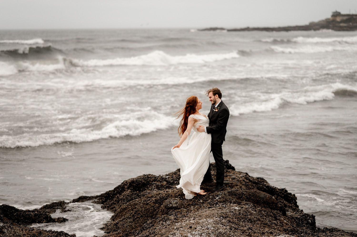 Bride and groom embrace on a rocky shore with ocean waves in the background at the Yachats Inn.