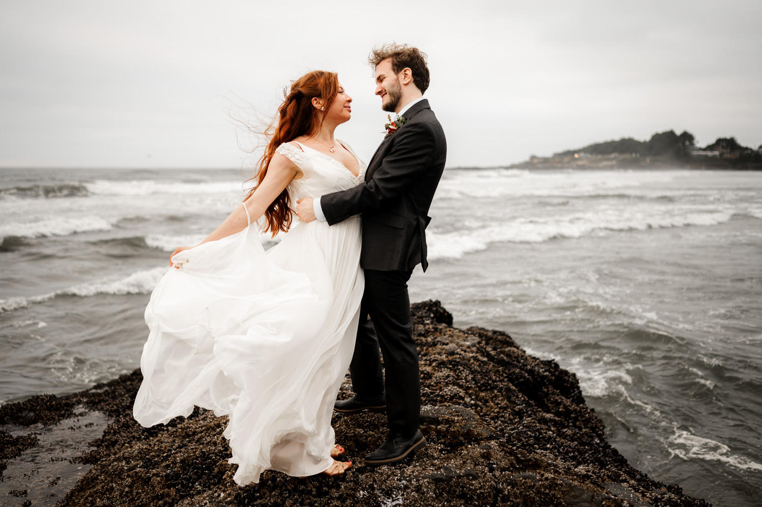 A bride and groom smiling at each other on a rocky shore, with waves in the background; wedding photos taken at the Yachats Inn.