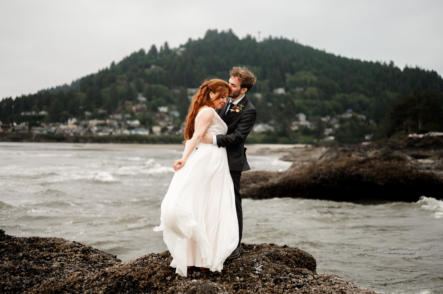 Bride and groom embrace on rocky shore with forested hills in background at Yachats Inn, capturing their wedding photos.