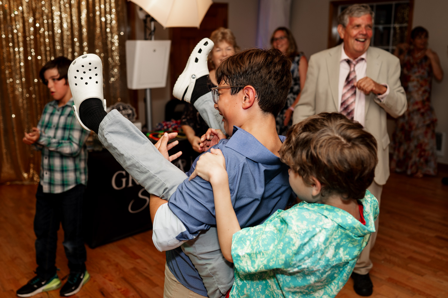 Two boys are playing joyfully at a party, with smiling adults in the background and a sparkling curtain behind them.