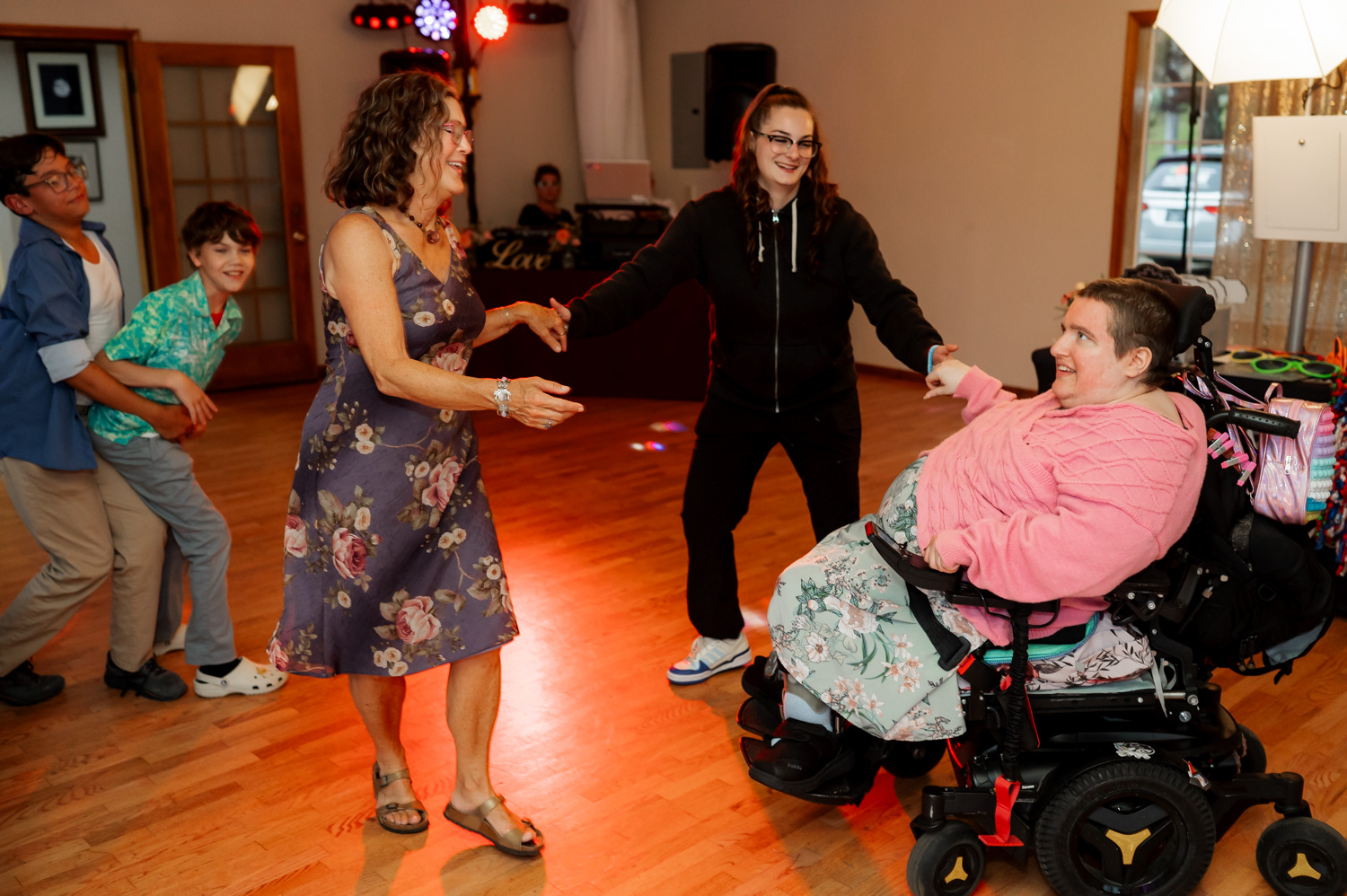 In a warmly lit room at the Yachats Inn, people are dancing energetically. One dancer is in a wheelchair, and there is a DJ booth in the background contributing to the lively atmosphere.